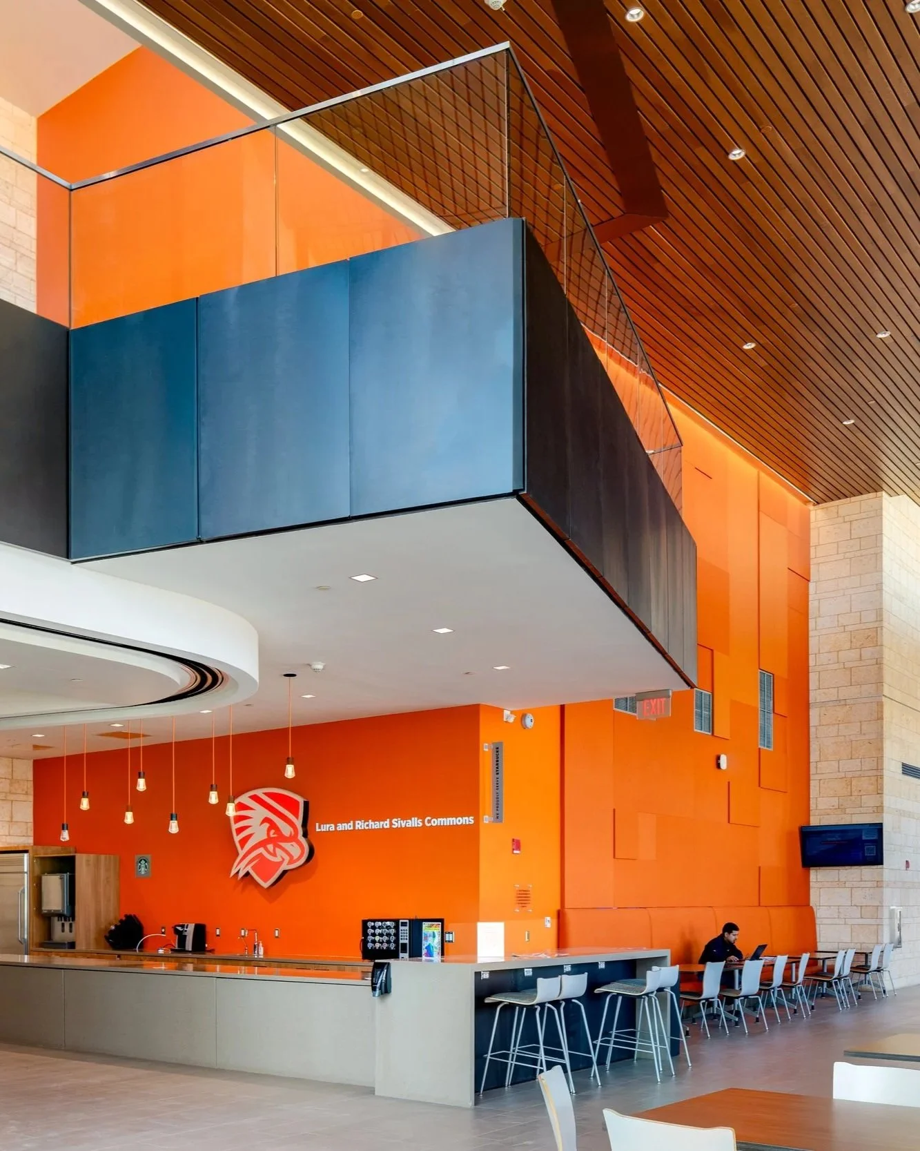 Interior of a modern lounge or cafeteria area with orange walls, a beige counter, bar stools, and a man using a laptop. A wall sign reads 'Lura and Richard Sivalls Commons' with a lion logo.