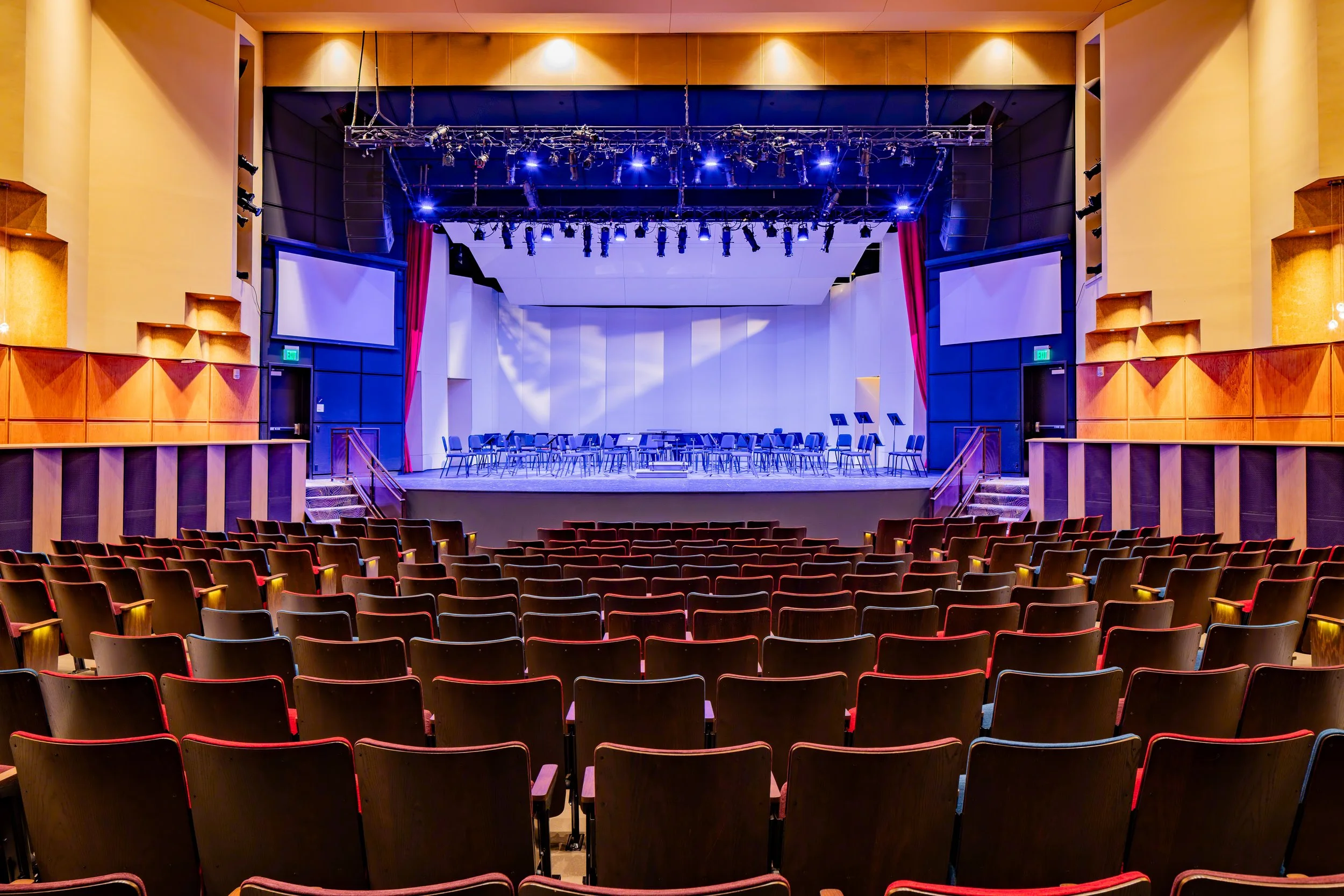 Empty auditorium theater with rows of chairs facing a stage with music stands and chairs, surrounded by wood paneling and equipped with stage lighting and large screens.