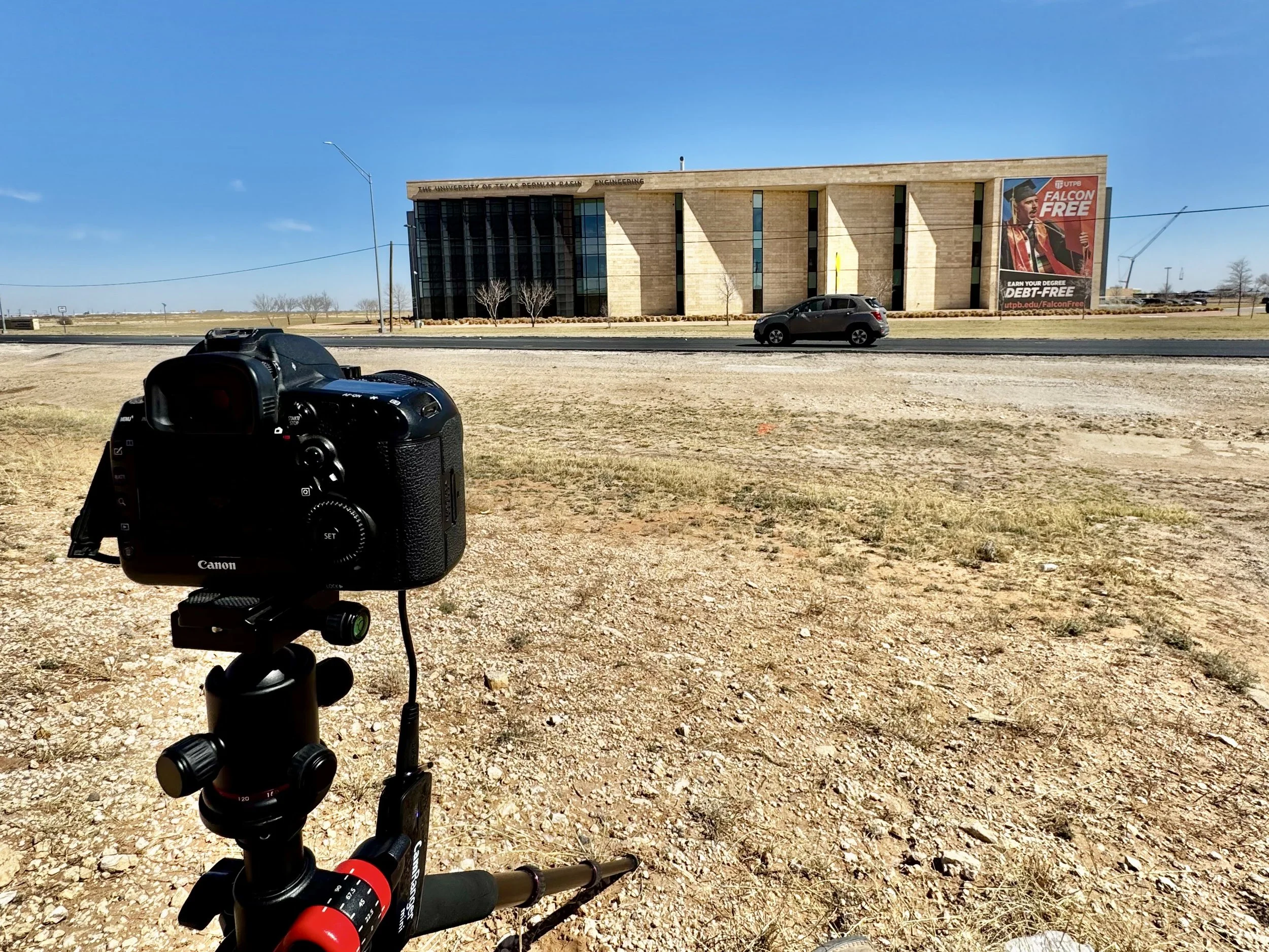 A Canon camera mounted on a tripod in an open, dry, grassy area facing a modern university building with a large billboard free advertisement for a degree program, under a clear blue sky.