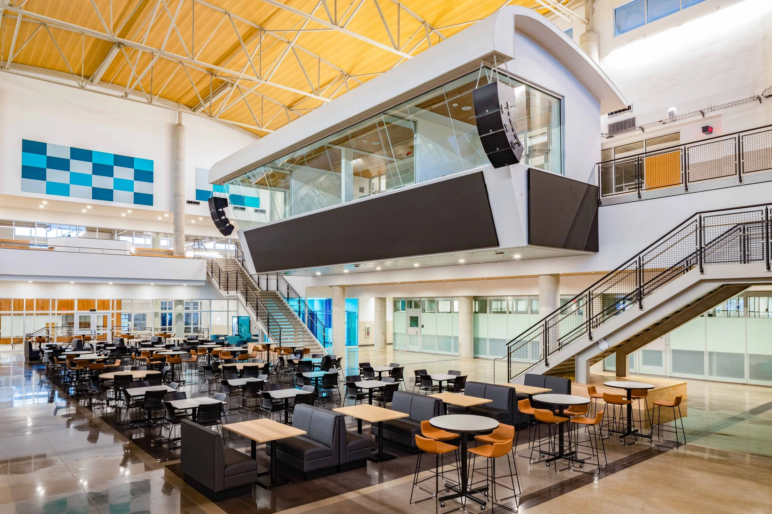 Interior of a modern, spacious building with a high wooden ceiling, large glass windows, and multiple levels connected by stairs. The lower level has numerous tables and chairs, and there are large black speakers hanging from the ceiling.