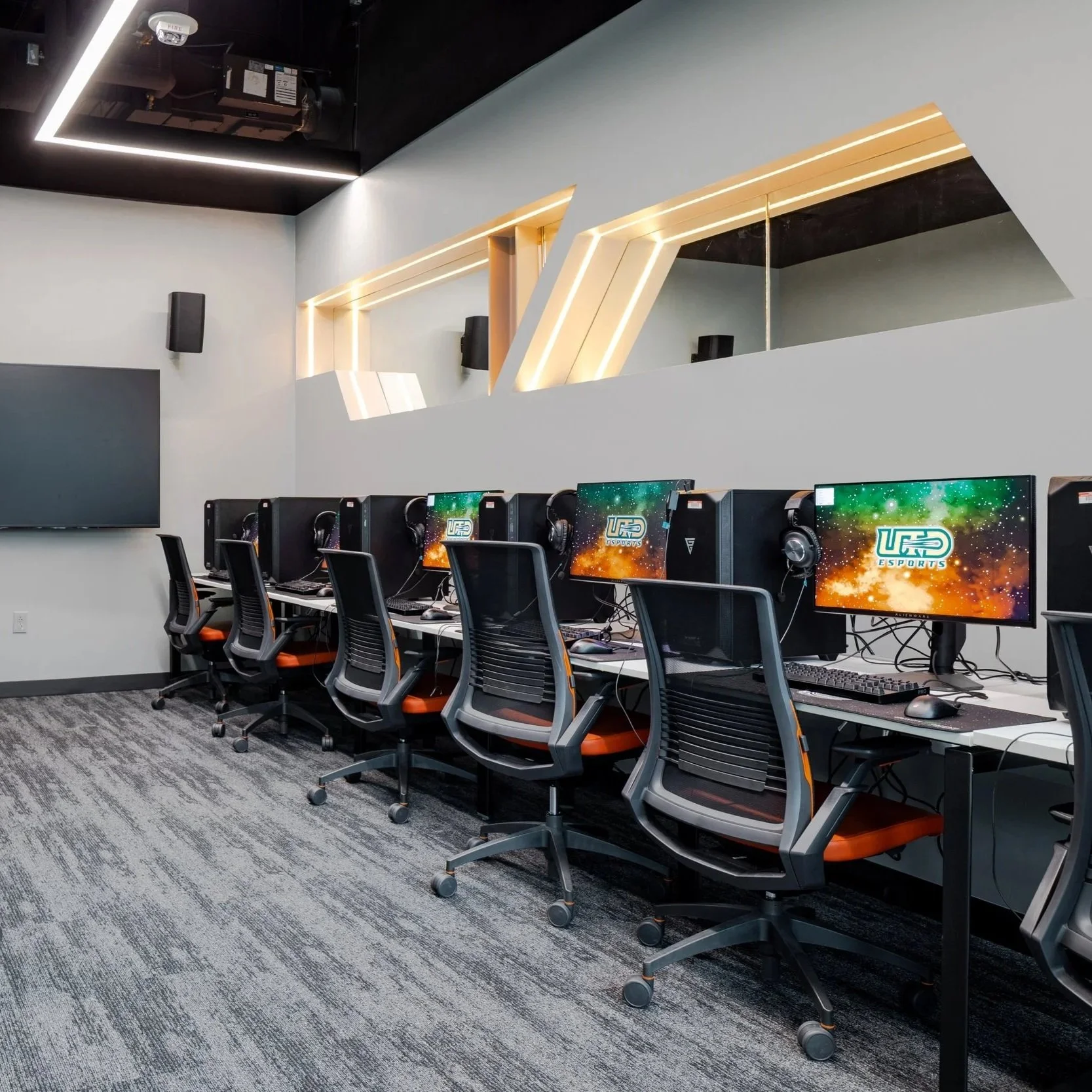 An esports gaming room with a row of gaming computers on a long table, each equipped with monitors, headphones, and gaming accessories. Black office chairs with orange accents are positioned in front of each computer. The room has geometric wall design with integrated lighting, black ceiling with visible ventilation and lighting, large wall-mounted flat-screen TV, and a dark patterned carpet.