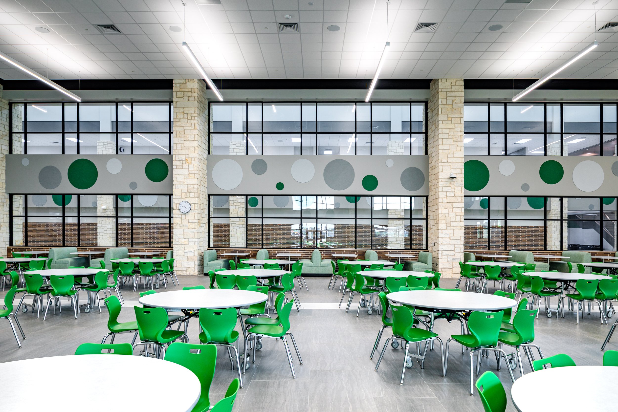 A modern cafeteria with green chairs and white round tables, large windows, stone columns, and a wall with circular green and gray patterns.
