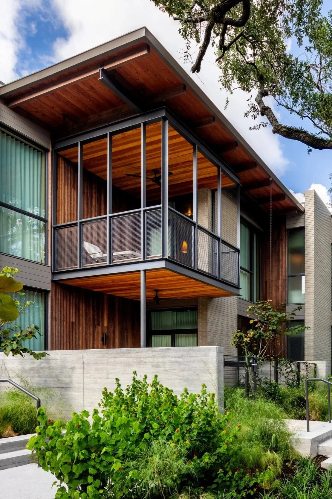 Modern residential building with large glass windows, wooden and brick exterior, a balcony with metal railing, and landscaped greenery in the foreground.