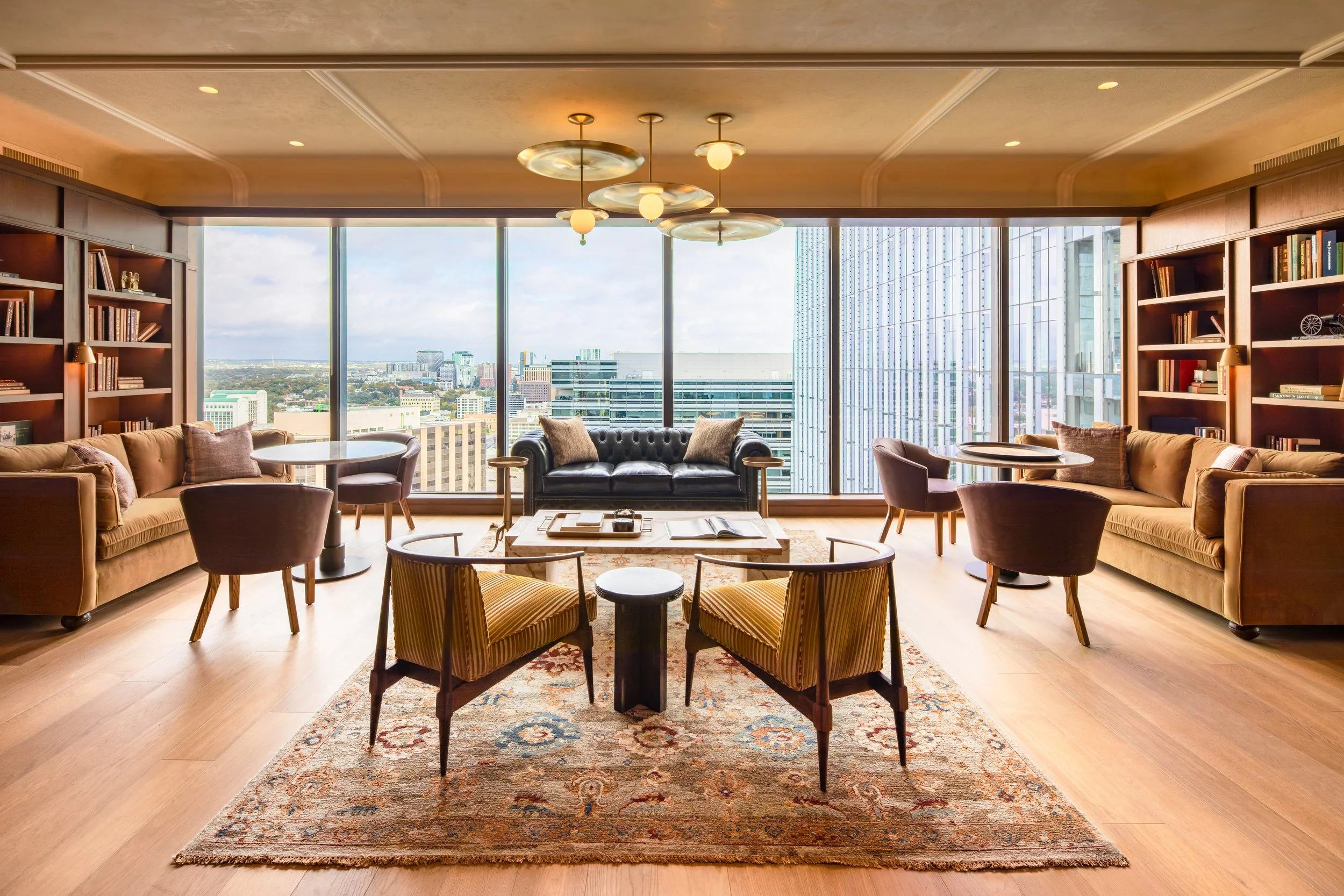 Sunlit lounge with large floor-to-ceiling windows showing cityscape, furnished with sofas, armchairs, a coffee table, bookshelves, and decorative lighting.
