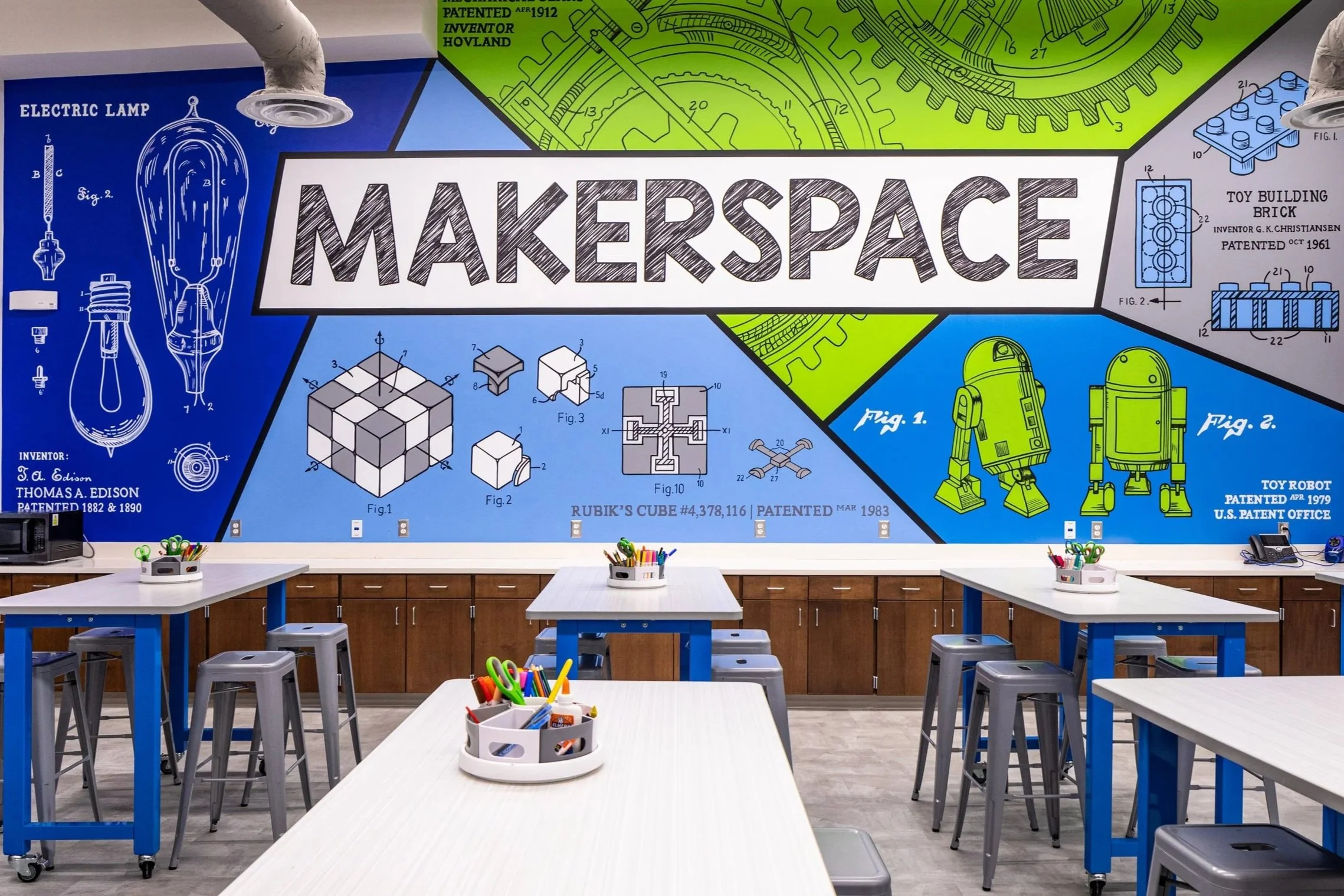 Colorful classroom with a large mural titled 'Makerspace' featuring diagrams of lightbulbs, Rubik's cube, toy robot, and mechanical gears, with classroom tables and art supplies.