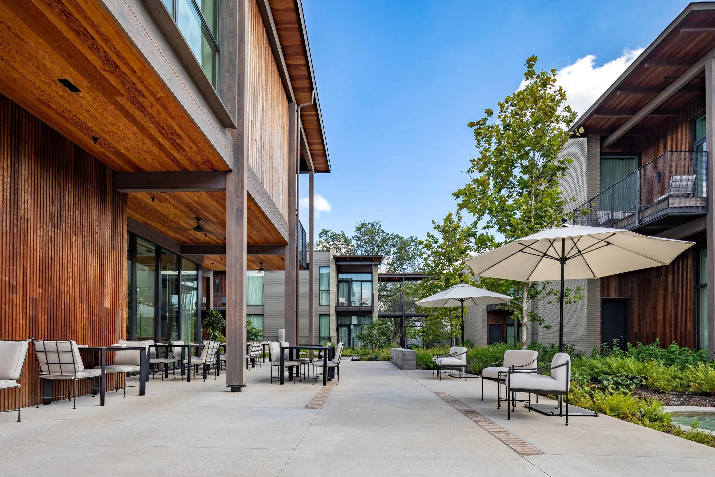 Modern apartment courtyard with outdoor seating, umbrellas, trees, and wooden building facades.