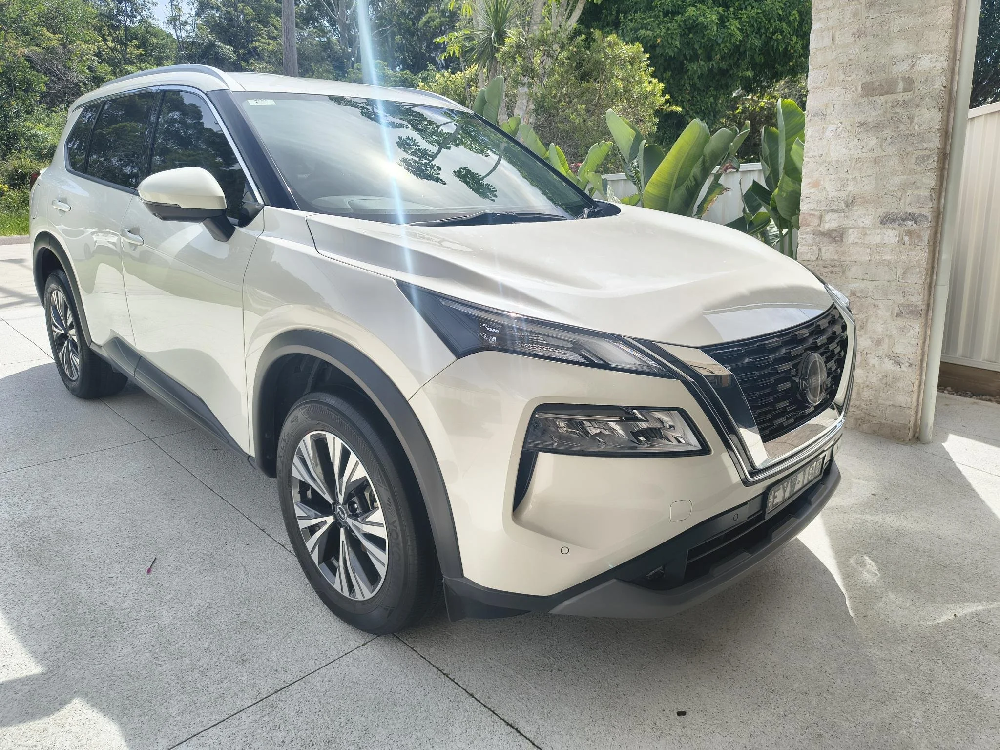 White Nissan SUV parked on a driveway next to a brick wall and green tropical plants, with sunlight reflecting off its windows.