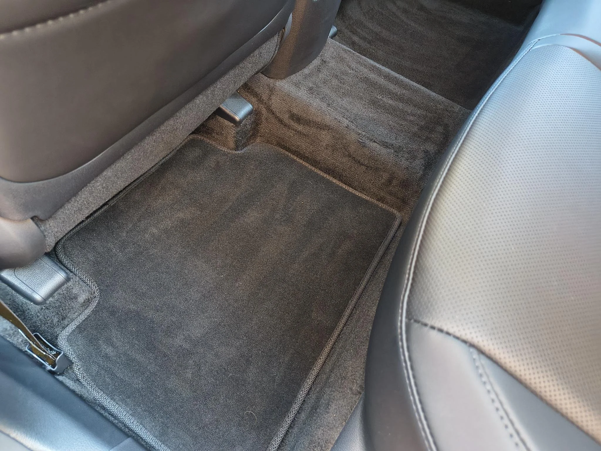 Car's back seat area showing a black floor mat on the floor with beige carpeting around it, and part of a light gray leather seat on the right.
