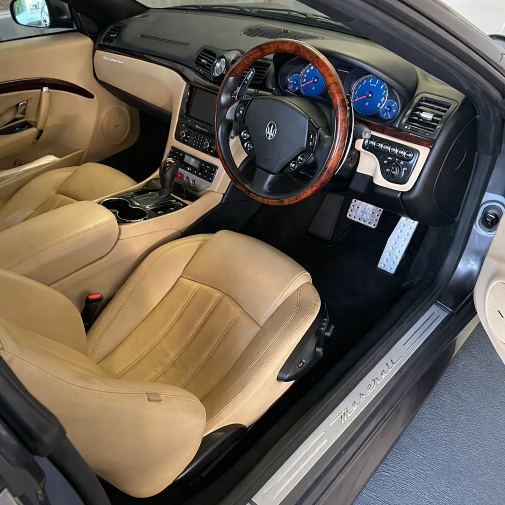 Interior of a luxury Maserati car with beige leather seats, a wood-trimmed steering wheel, and a dashboard with blue instrument gauges.