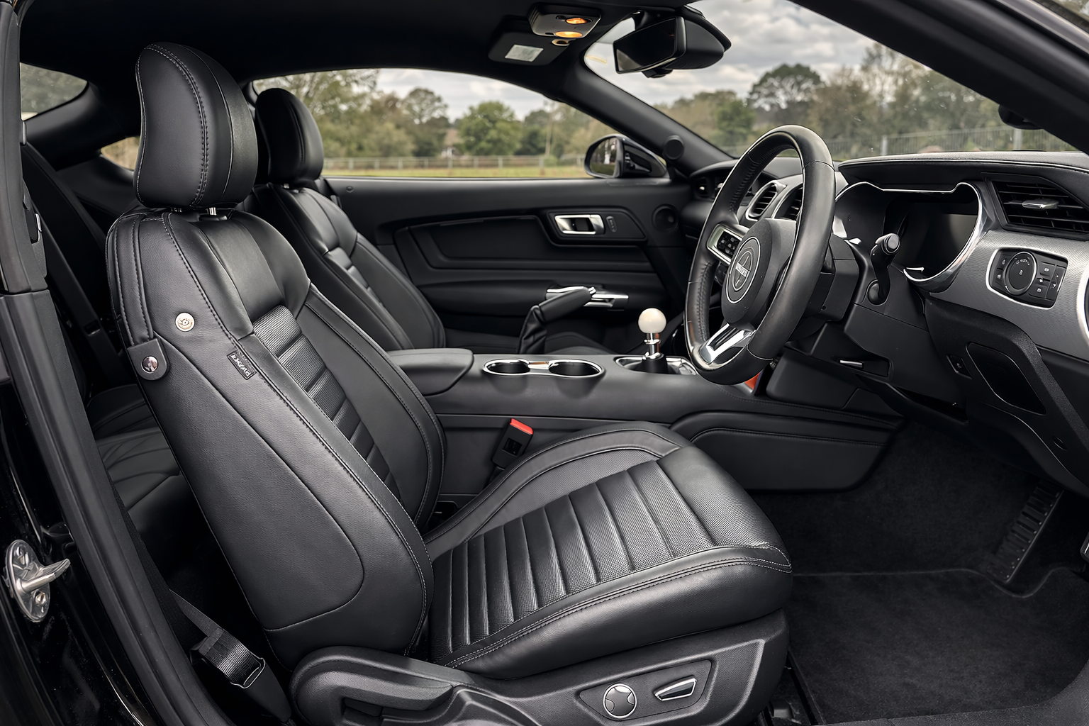 Inside a modern vehicle with black leather seats, a steering wheel, gear shifter, and dashboard, with a scenic outdoor view through the windshield.