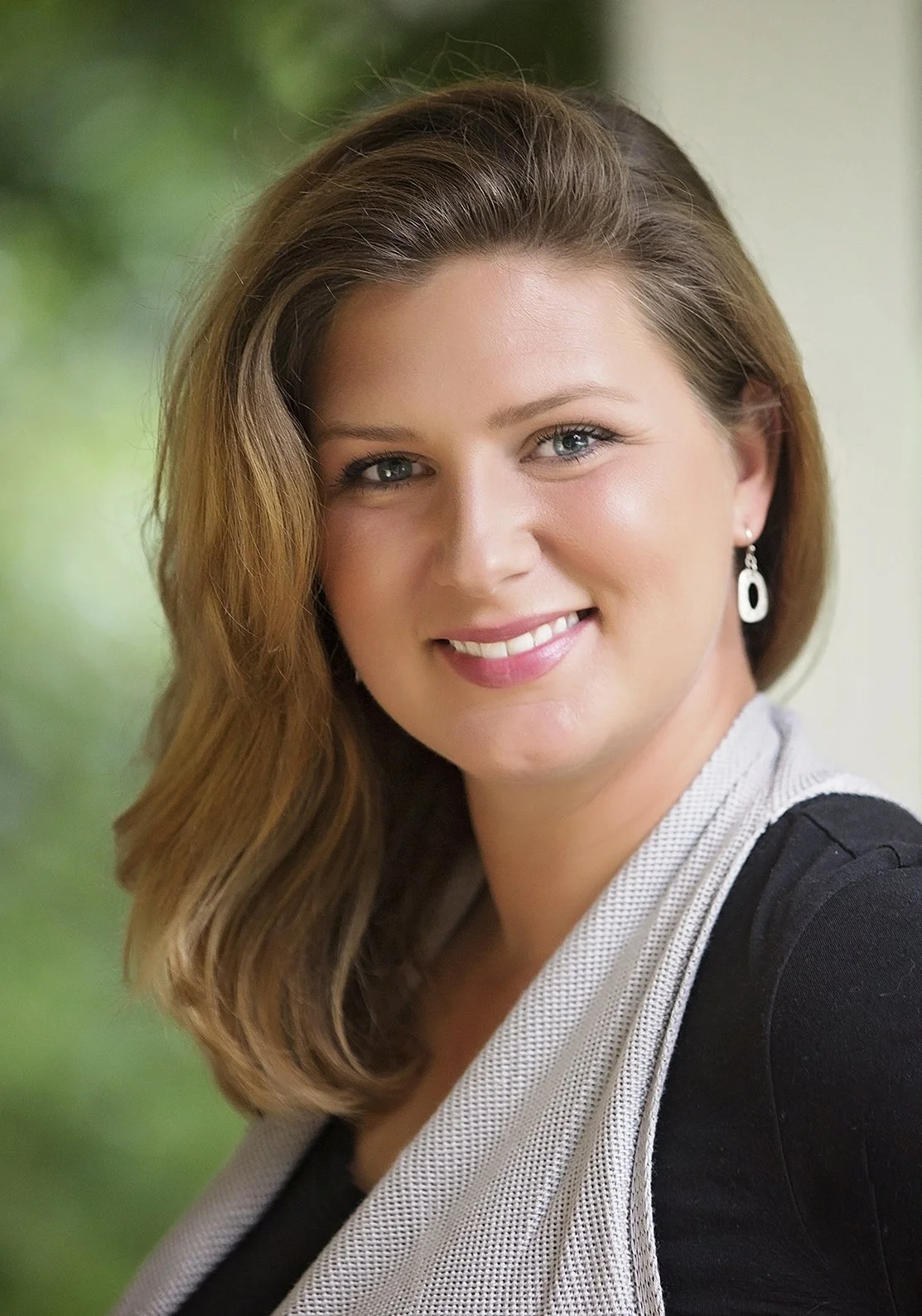 Close-up of a woman with shoulder-length light brown hair, blue eyes, smiling, wearing a black top, a light-colored scarf or vest, and earrings, with a blurred green background.