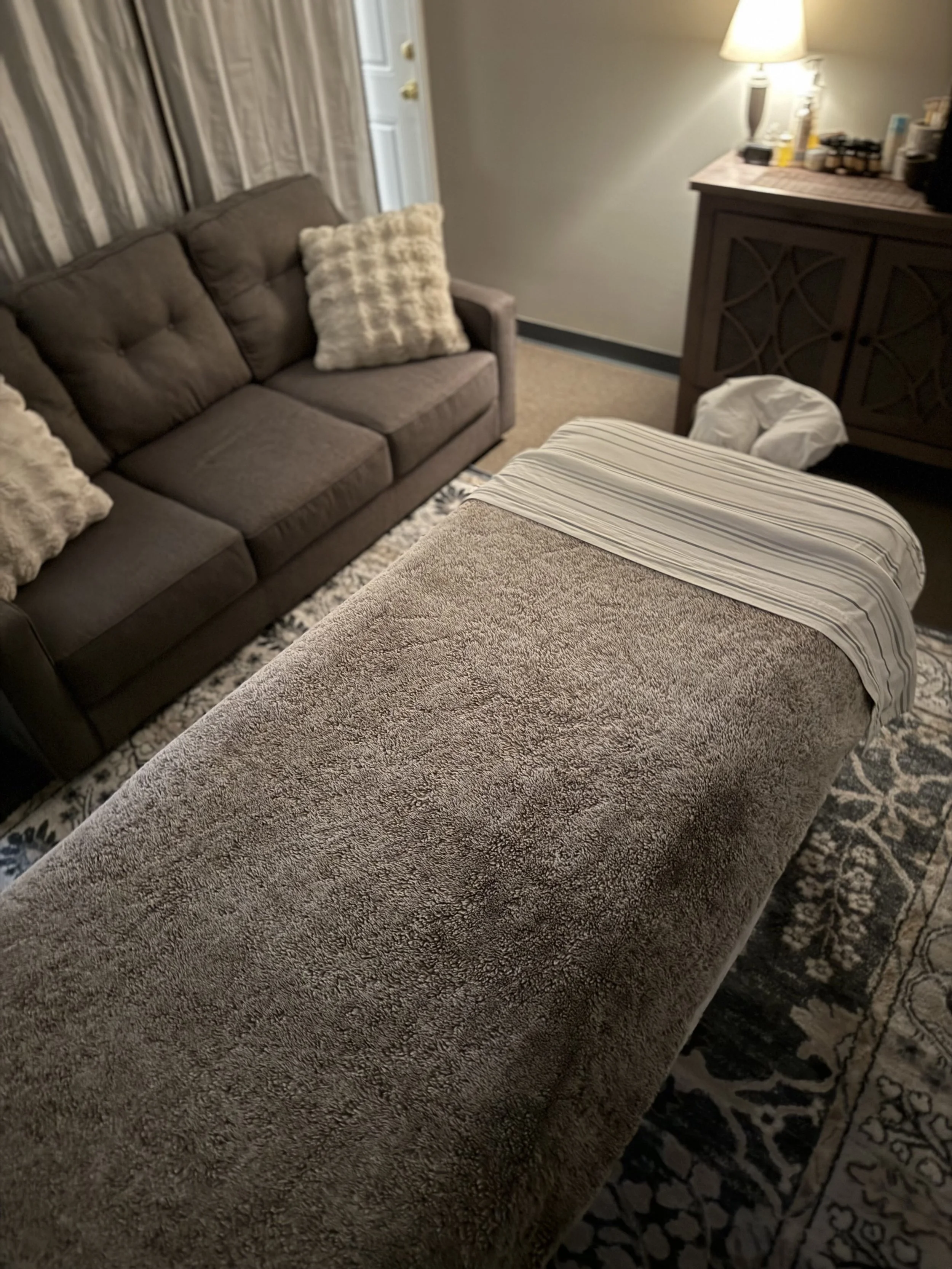 A massage table with a fuzzy grey blanket and a striped sheet in a cozy room. In the background, a dark grey sofa with fur pillows, a side table with a lamp, and a cabinet with miscellaneous items on top.