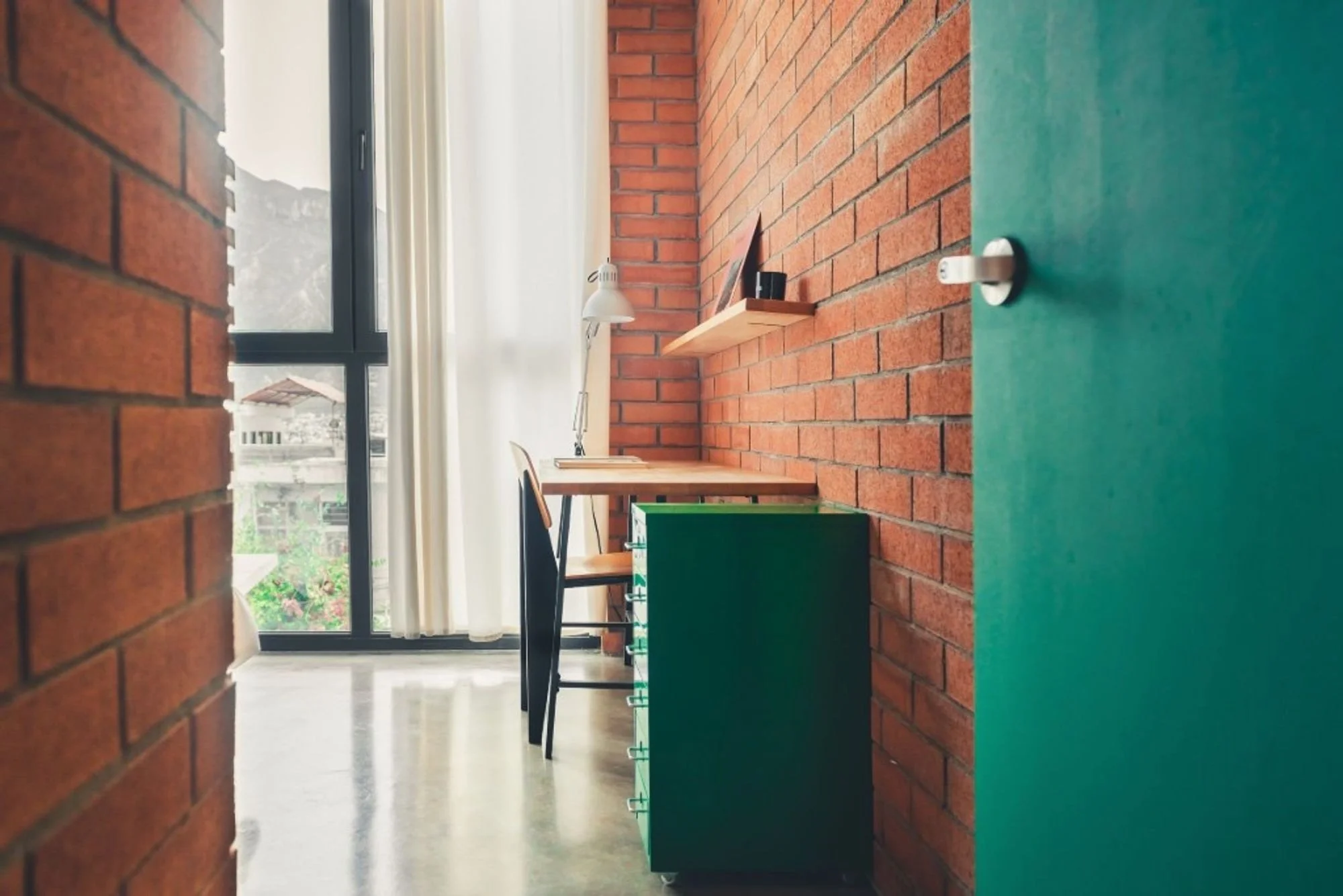 View of a bright room with brick walls, a large window with curtains, a desk with a lamp, chairs, and a green cabinet, seen through a partially open door.