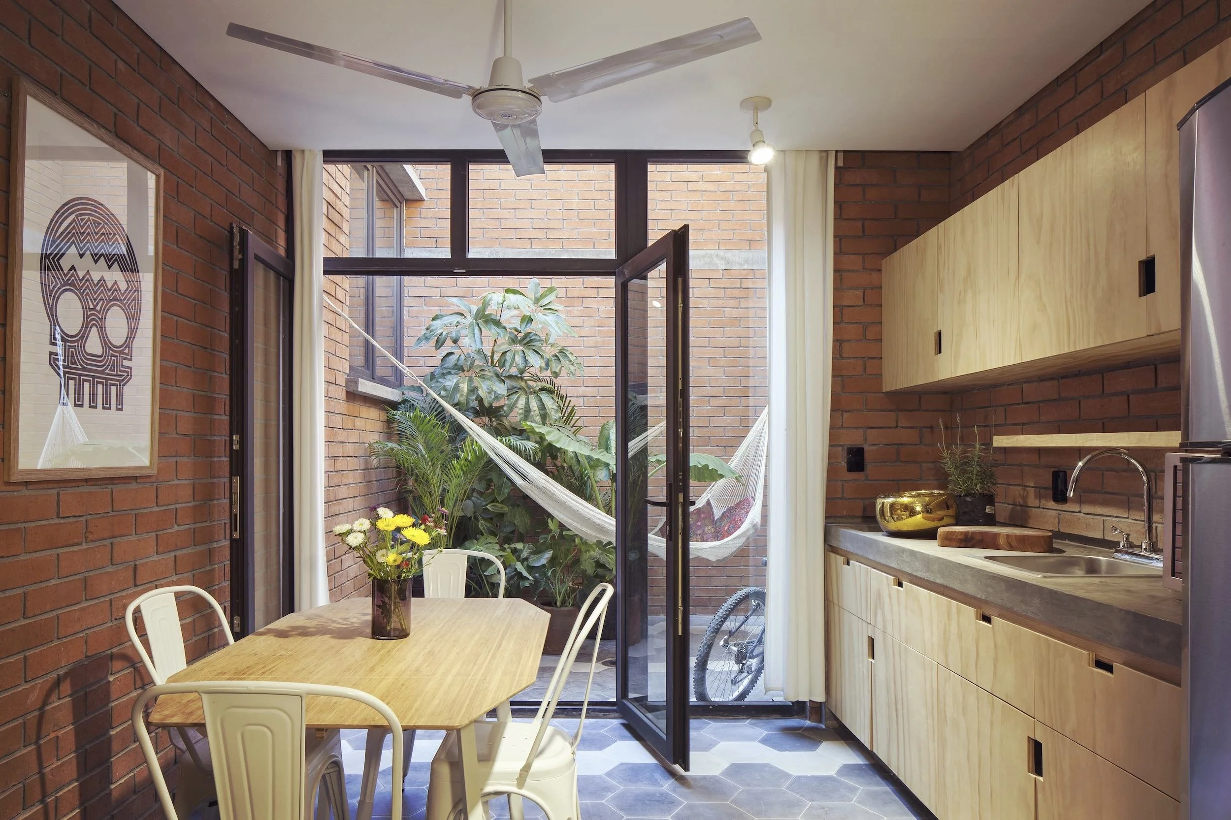 Kitchen area with brick walls, a wooden table with white chairs, a vase of flowers, open glass door leading to a patio with greenery and a hammock, ceiling fan, and wooden cabinets.