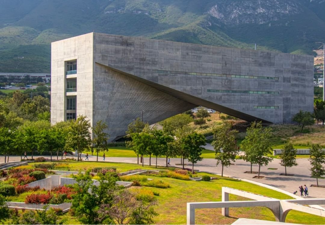 Modern concrete building with an angular design, surrounded by trees and landscaping, with mountains in the background.