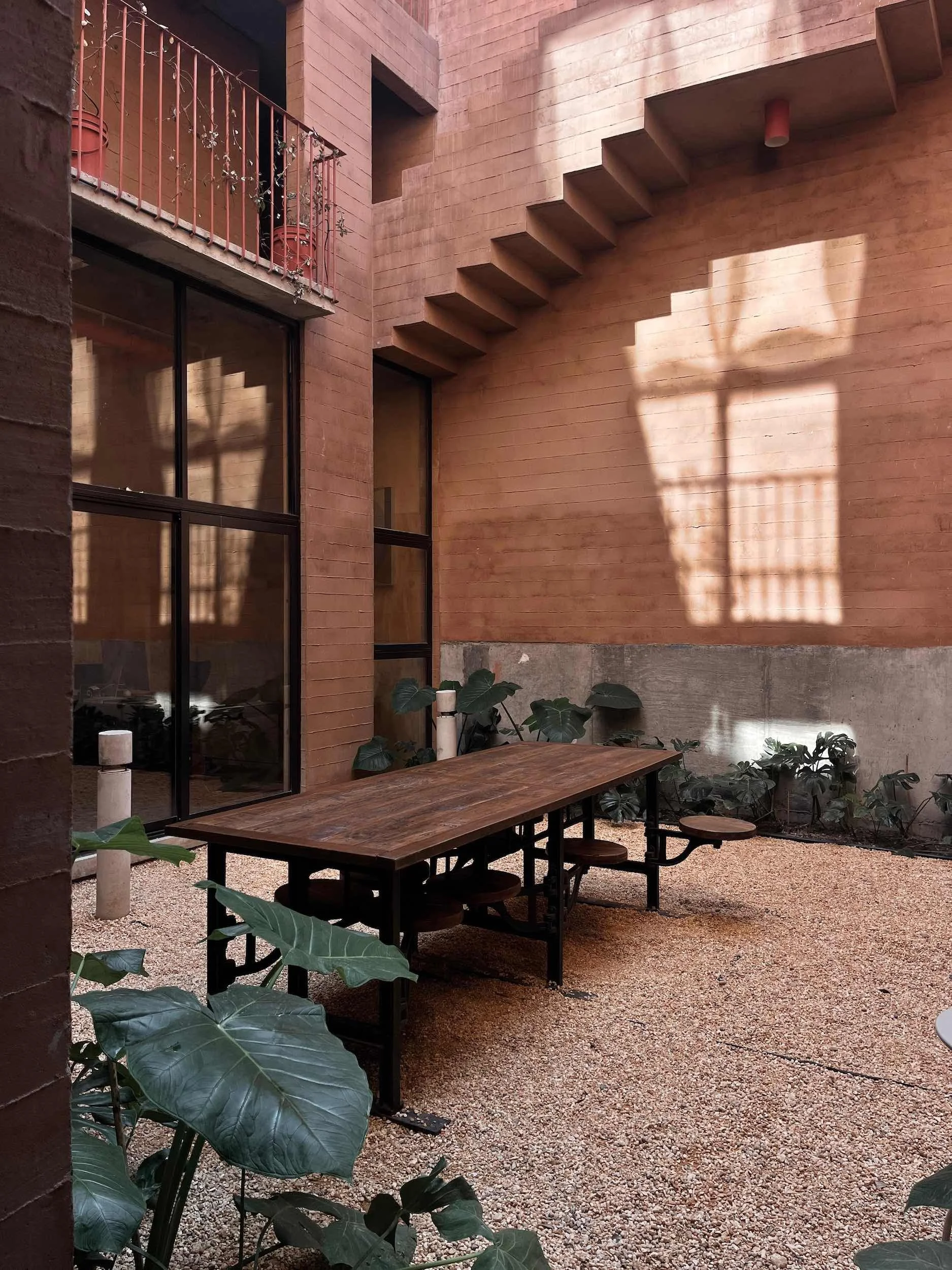 Outdoor courtyard with a wooden table and attached benches, surrounded by green plants, brick and concrete walls, and a staircase casting a shadow on the wall.