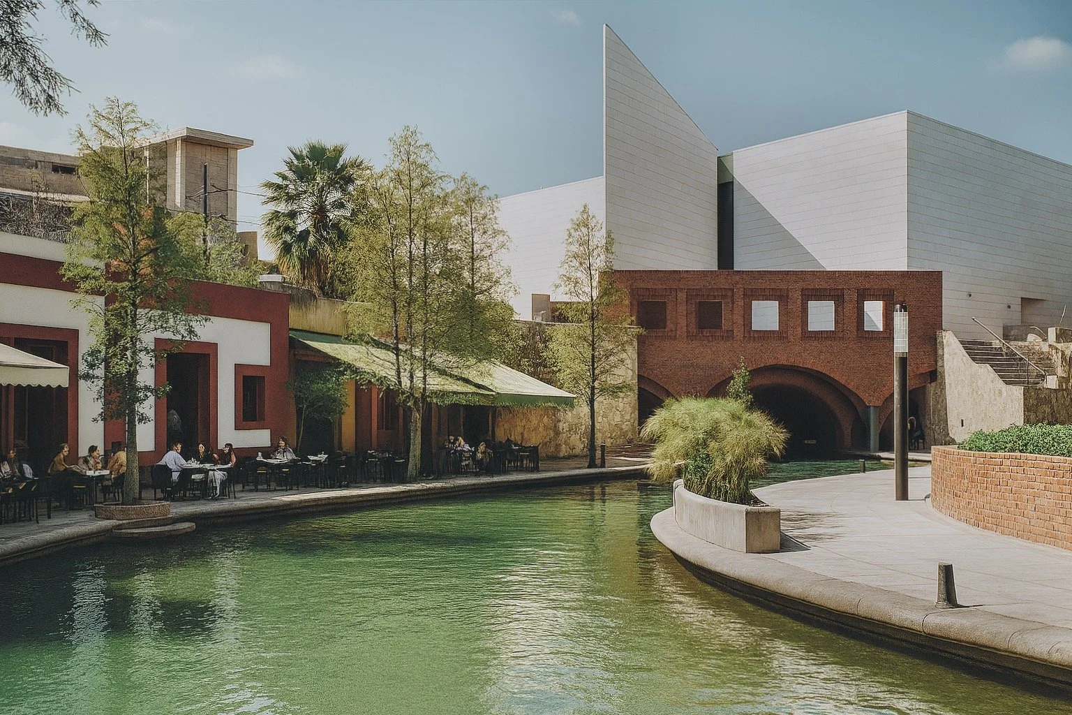 Outdoor scene with a river or canal, surrounded by trees, buildings, and a sidewalk with people dining at tables.