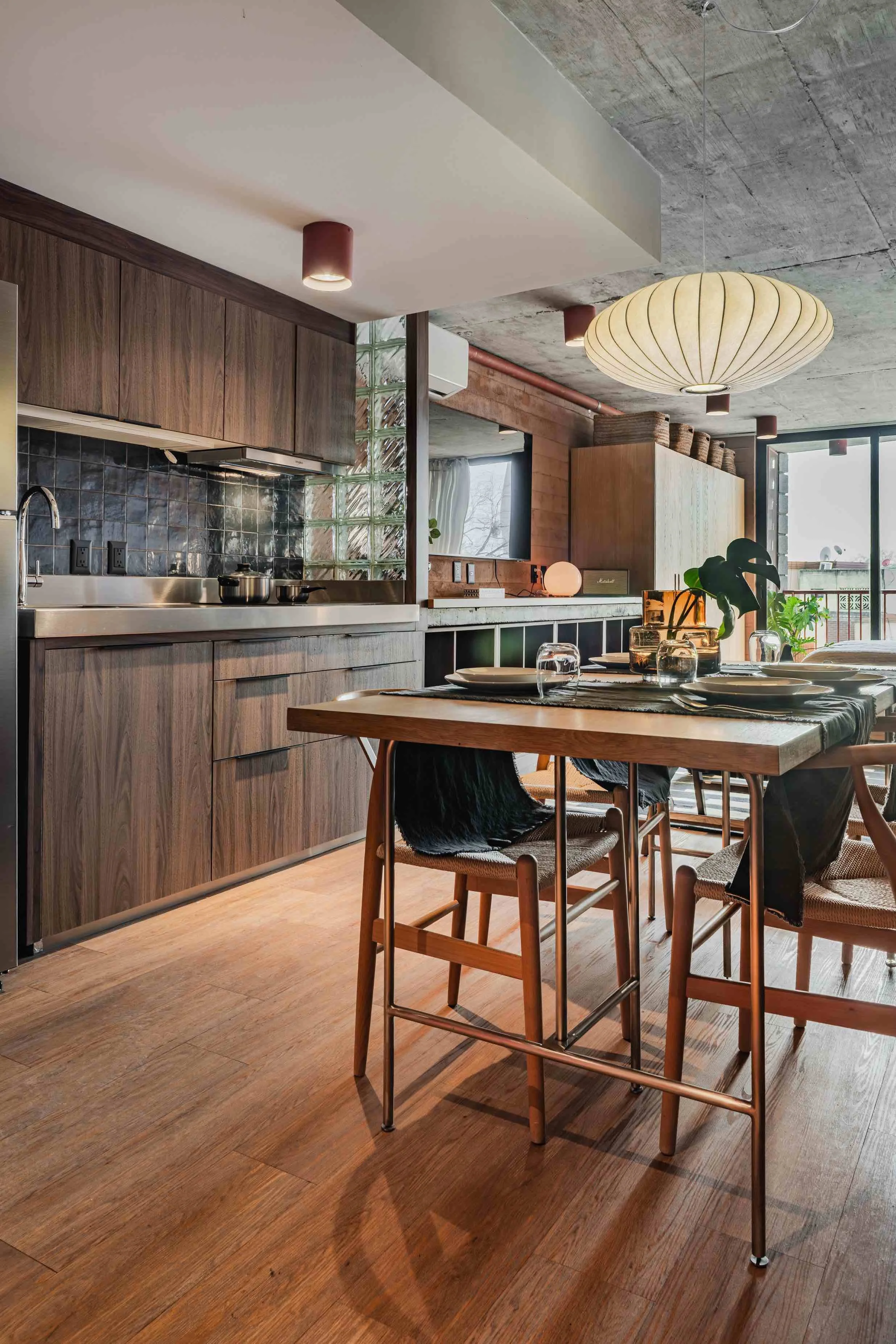 Modern kitchen and dining area with wooden cabinets, a wooden dining table set for four, hanging pendant lights, a large window, and decorative plants.