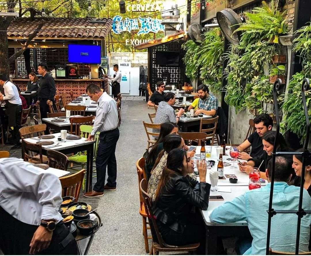 A lively outdoor restaurant or cafe with people dining and socializing. There are several tables with chairs, some people using their phones. Waitstaff are serving food. The setting is decorated with lush green plants and outdoor lights, creating a c