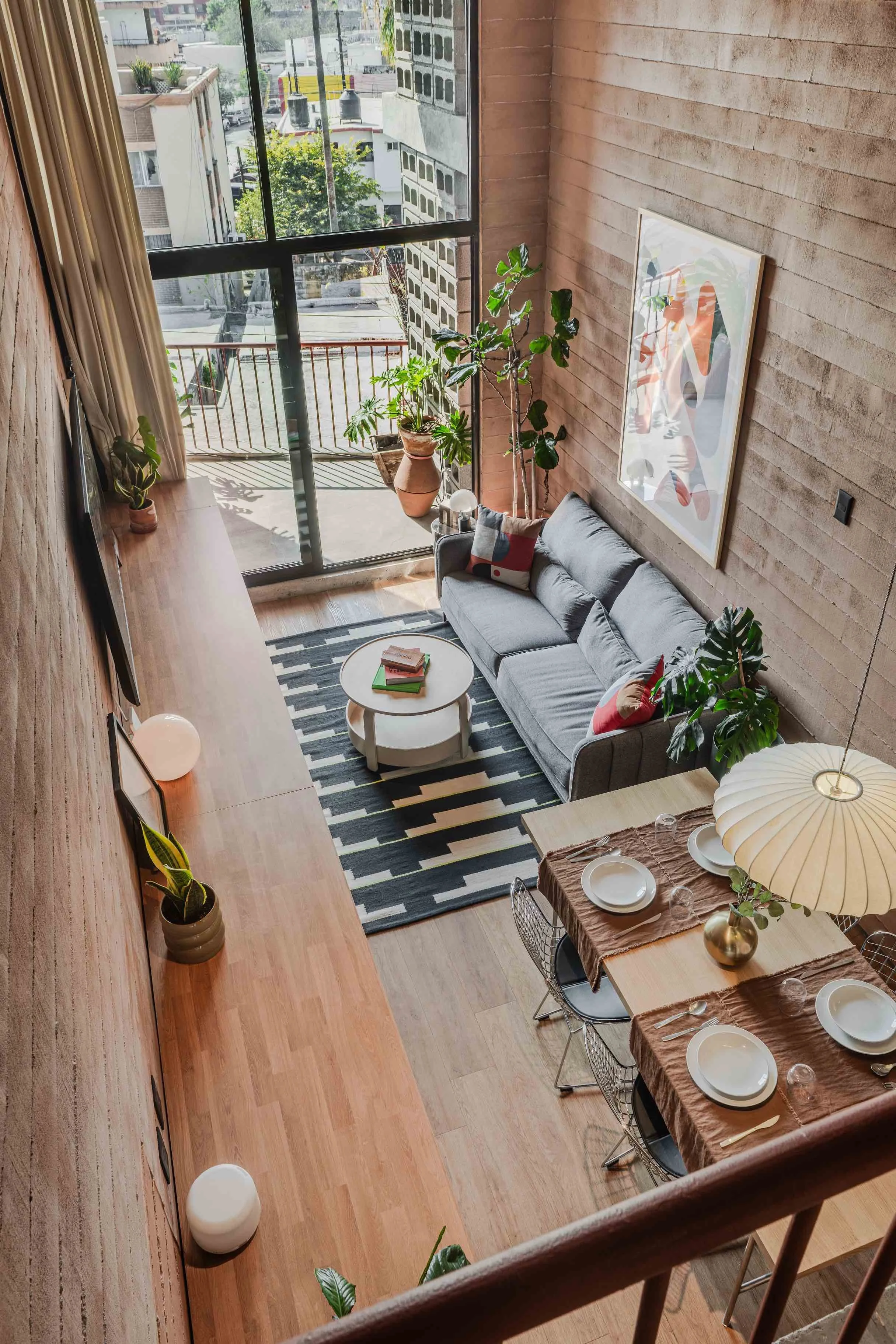 A modern living room with wooden flooring and a large window overlooking a city street. The room has a gray sofa with colorful pillows, a round coffee table with books, a striped rug, and indoor plants. A dining area with a table set with plates and 