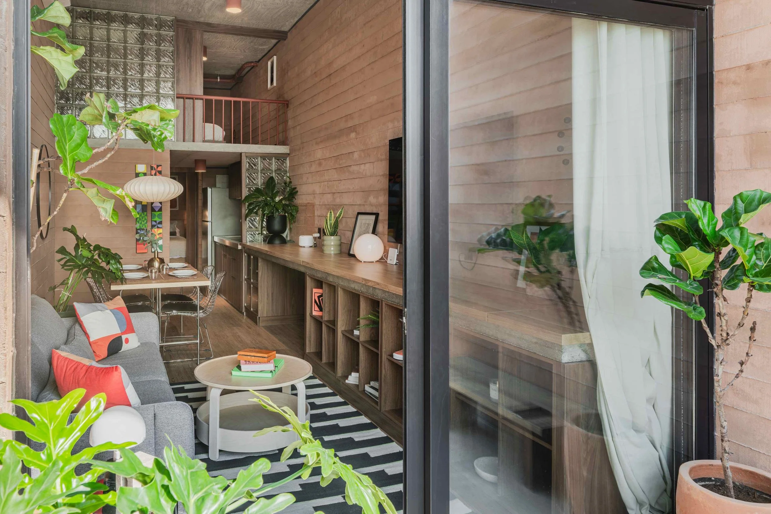 Interior of a modern living space with wooden walls, a gray sofa with colorful pillows, a round coffee table with books, and lush green plants. A dining area with a table and black chairs is visible in the background, along with decorative items and 