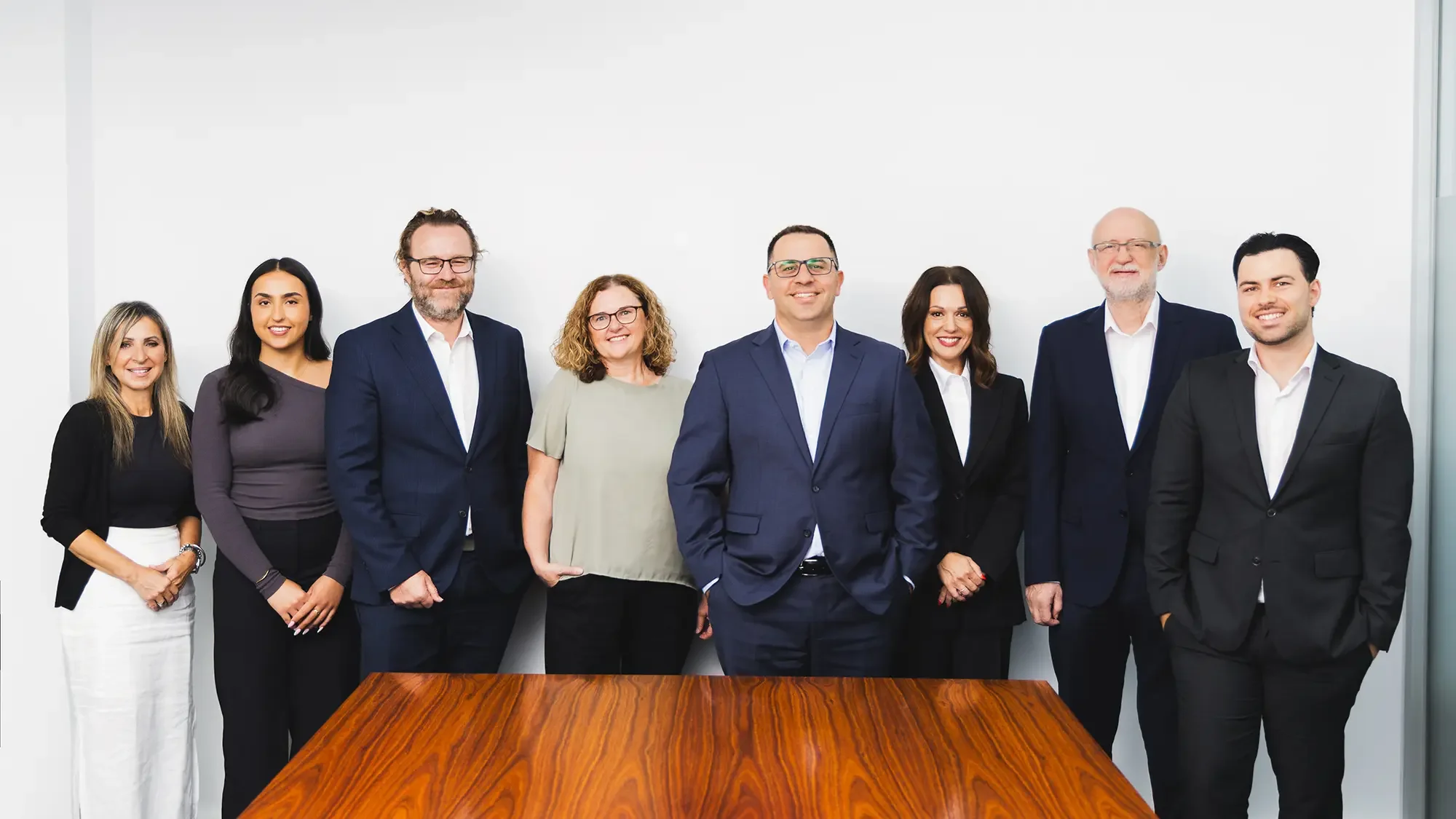 Group of eight diverse professionals standing in a conference room with a wooden table in the foreground, smiling at the camera.