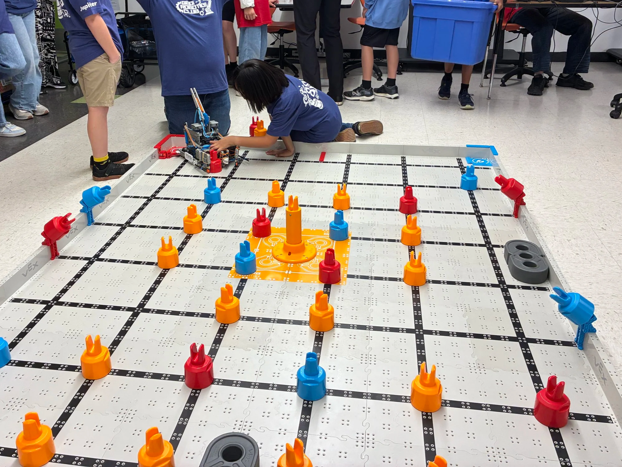 A group of children and adults gathered around a robotics project on a table, with a girl working on a robot in the center. The table has a grid pattern and colorful cone-shaped pieces.