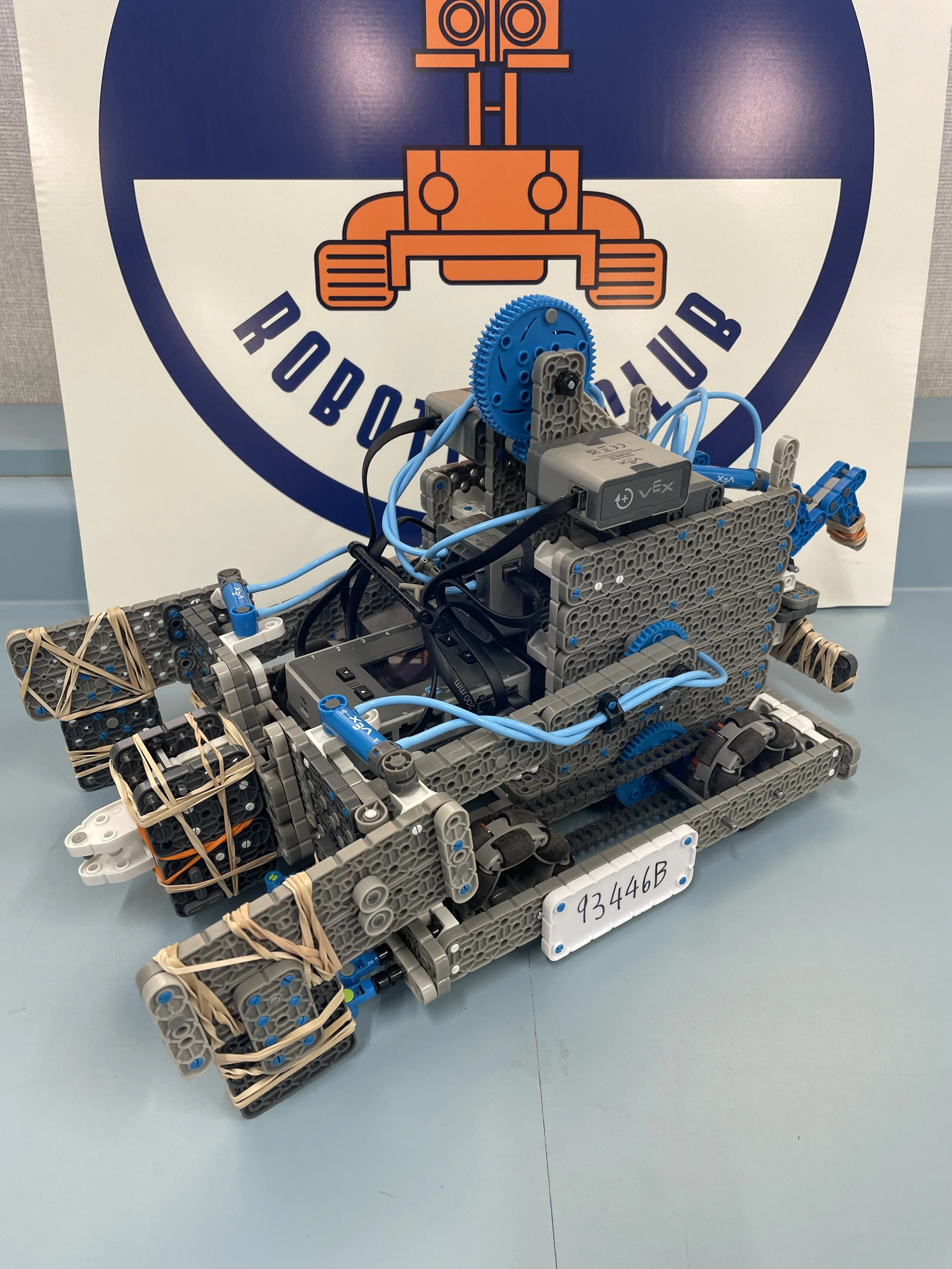 A LEGO robot with various gears, wires, and structural elements, displayed in front of a sign with a stylized robot logo and the text 'ROBOTICS CLUB.'