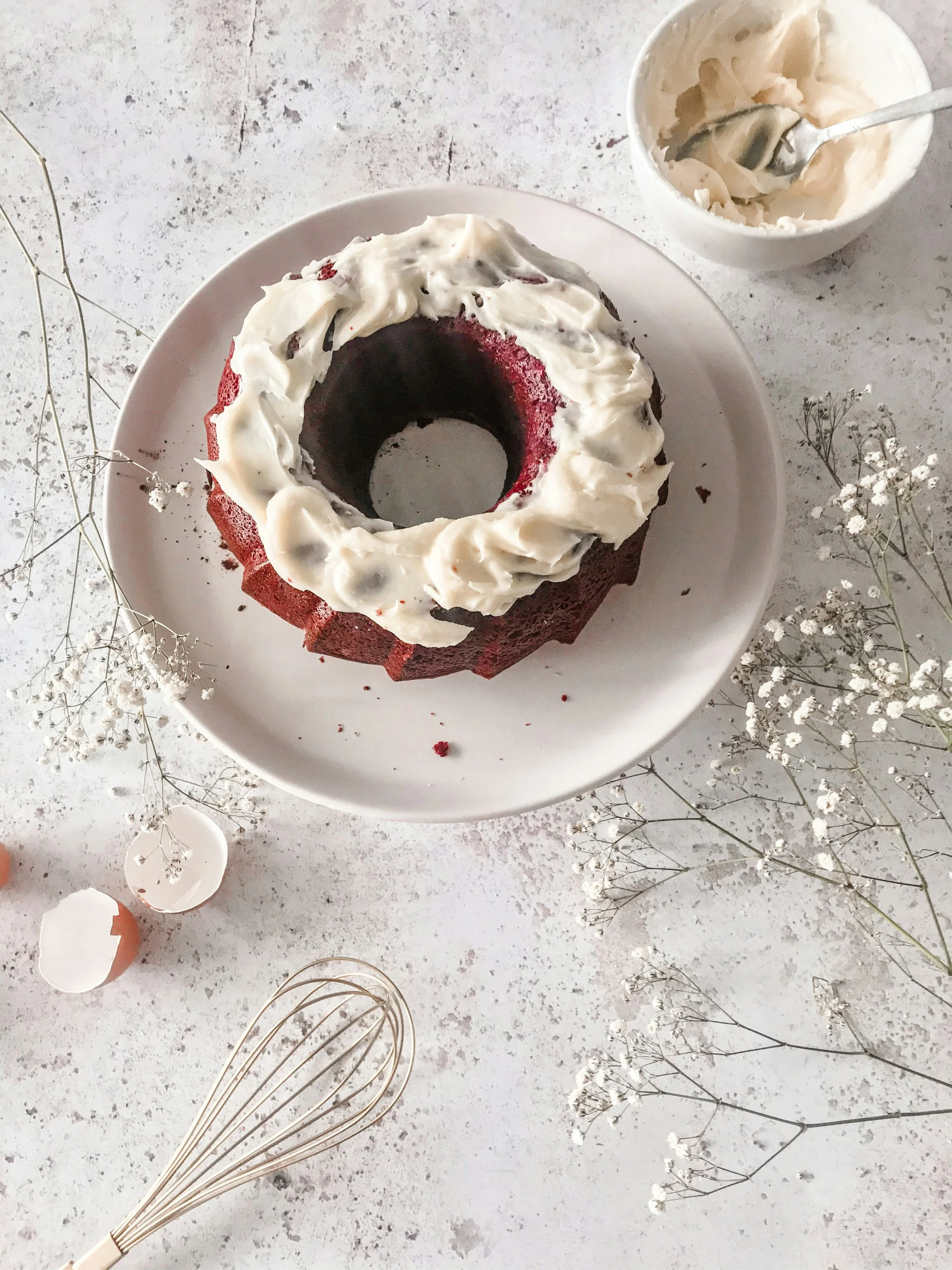 Red velvet Bundt cake topped with cream cheese frosting on a white plate, surrounded by eggshells, a whisk, and dried flowers.