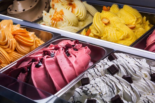 Assorted flavors of ice cream in a display freezer with pink, white, yellow, and tan varieties, some topped with fruit, nuts, or chocolate.