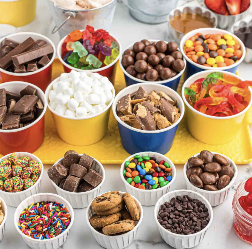 A colorful variety of candies and chocolates in small cups on a table, including chocolate bars, gummy bears, chocolate-covered nuts, jelly beans, marshmallows, sprinkles, chocolate chunks, cookies, chocolate chips, and Sour Patch Kids.