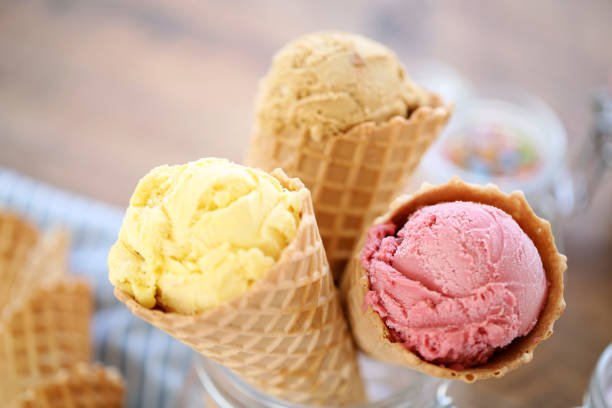 Three ice cream cones with scoops of yellow, beige, and pink ice cream on a wooden surface.