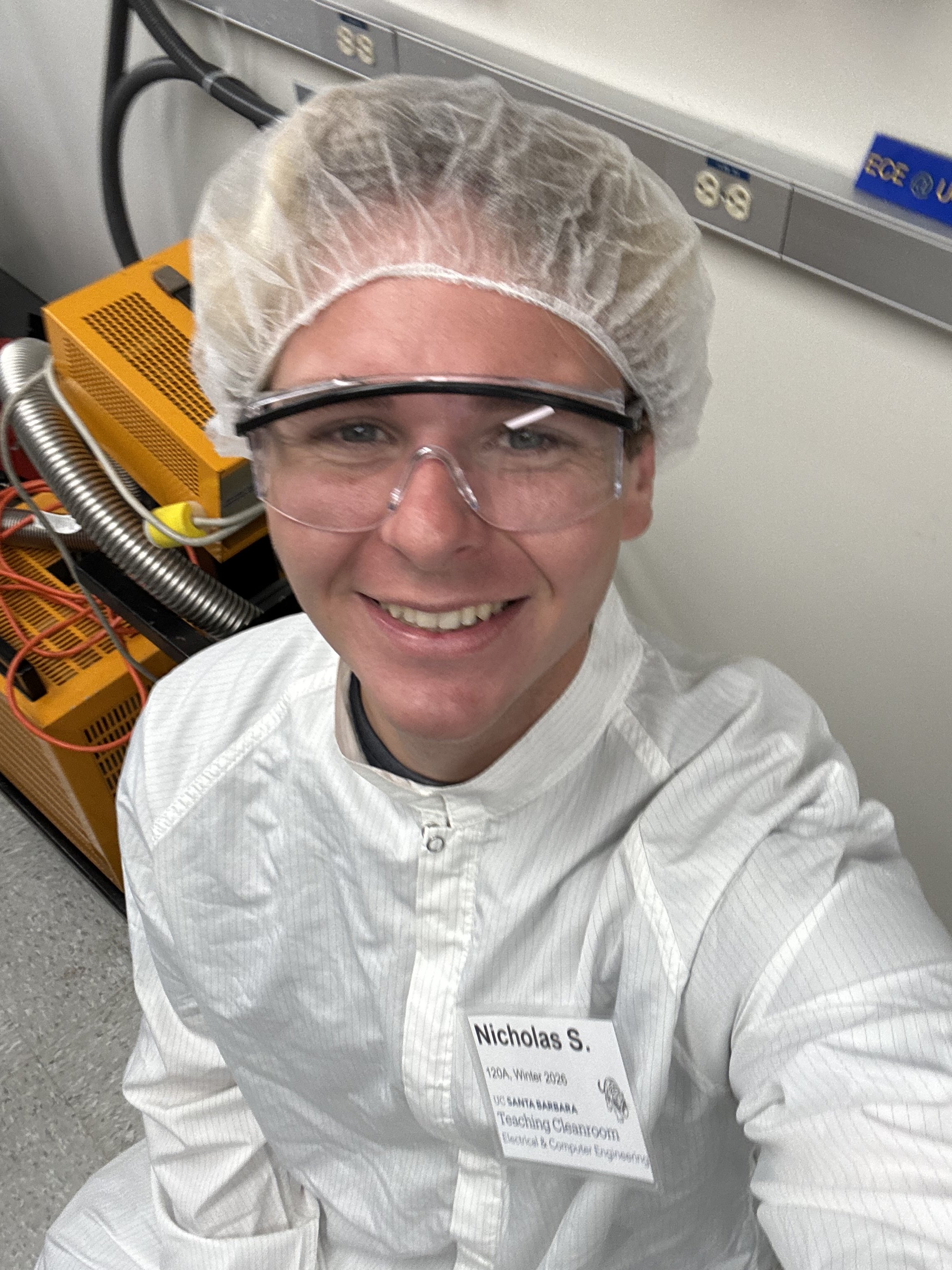 A person in protective gear, including safety goggles, a hair cover, and a white lab coat, smiling at the camera in a laboratory or cleanroom setting.
