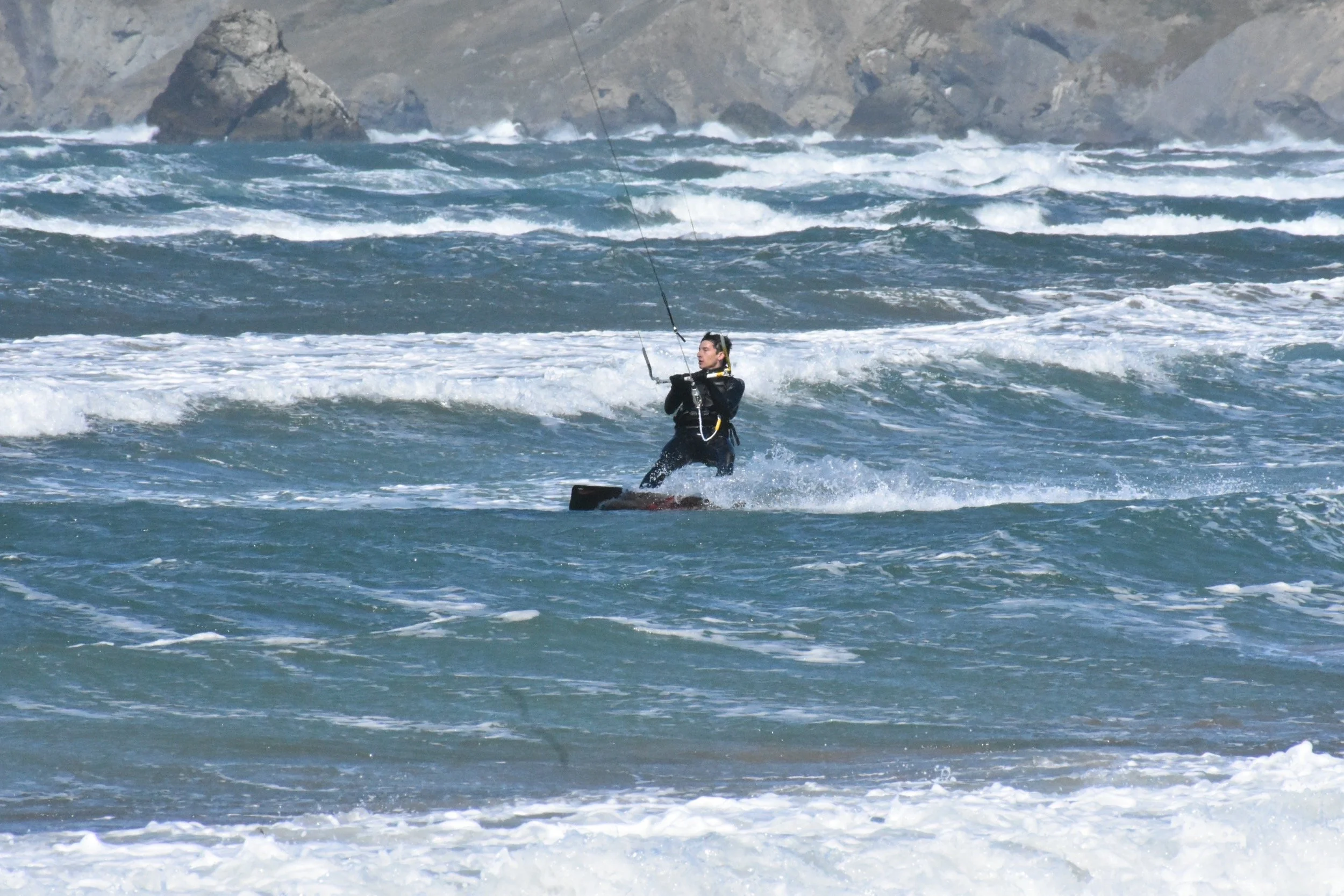 Person kiteboarding on ocean waves near rocky shoreline.