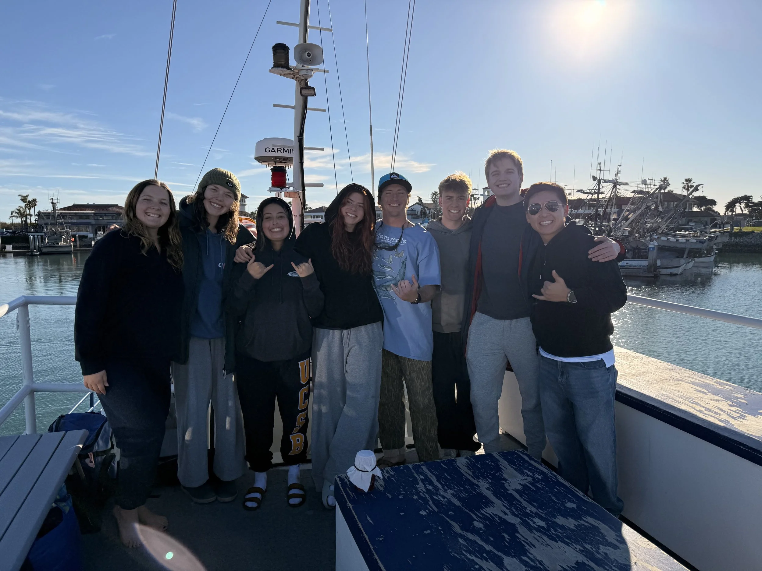 Group of nine friends standing on a boat dock, smiling at the camera, with boats docked in the background on a sunny day.