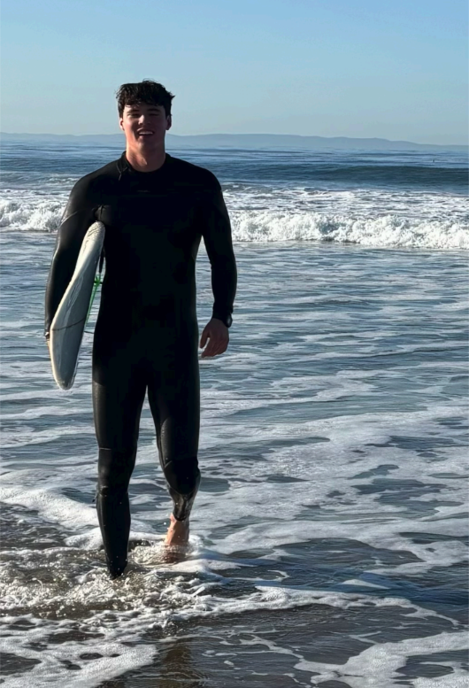 A young man standing in the ocean waves holding a surfboard, dressed in a black wetsuit at the beach on a sunny day.