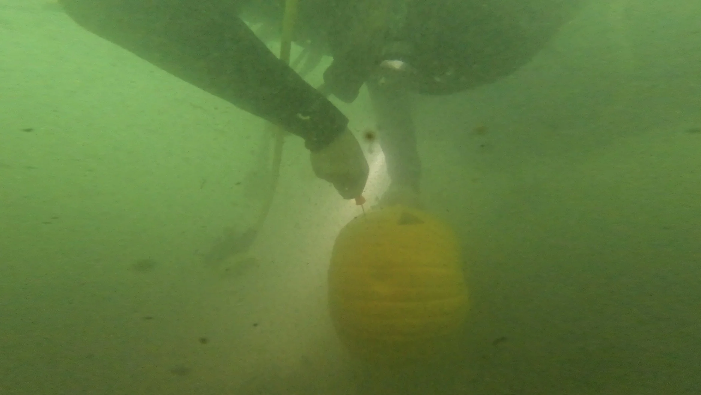 Underwater view of a yellow buoy hanging beneath a boat with greenish water.