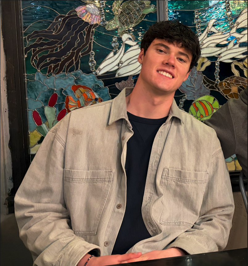 A young man with dark hair smiling, wearing a beige jacket over a black shirt, sitting indoors in front of a colorful stained glass mosaic underwater scene with fish, coral, and sea turtles.