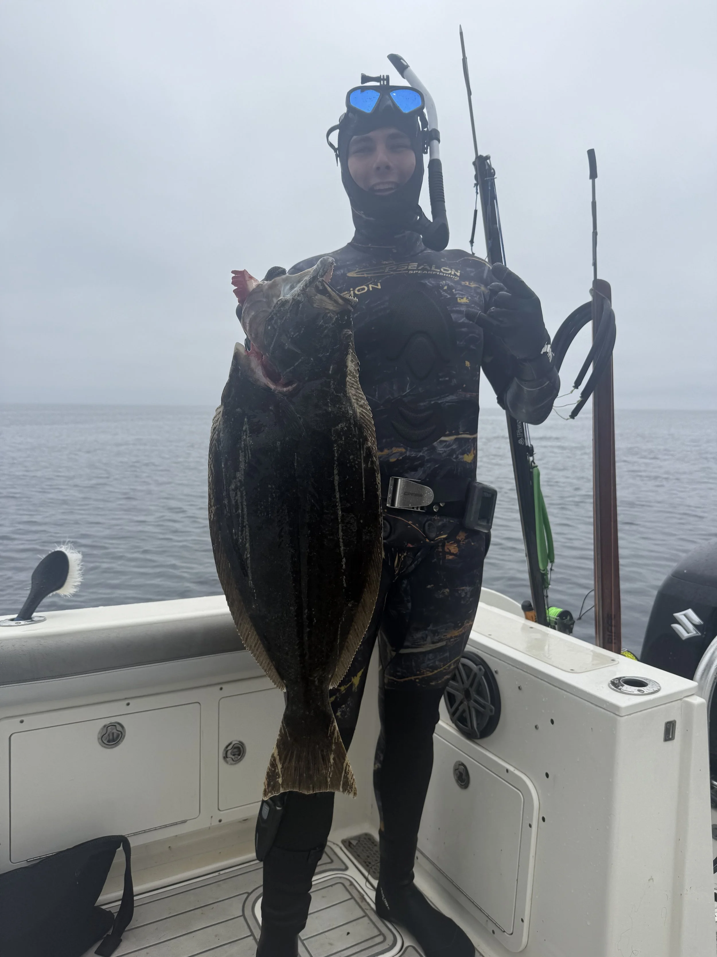 A person dressed in scuba diving gear holding a large flatfish on a boat at sea, with fishing rods in the background and cloudy sky.