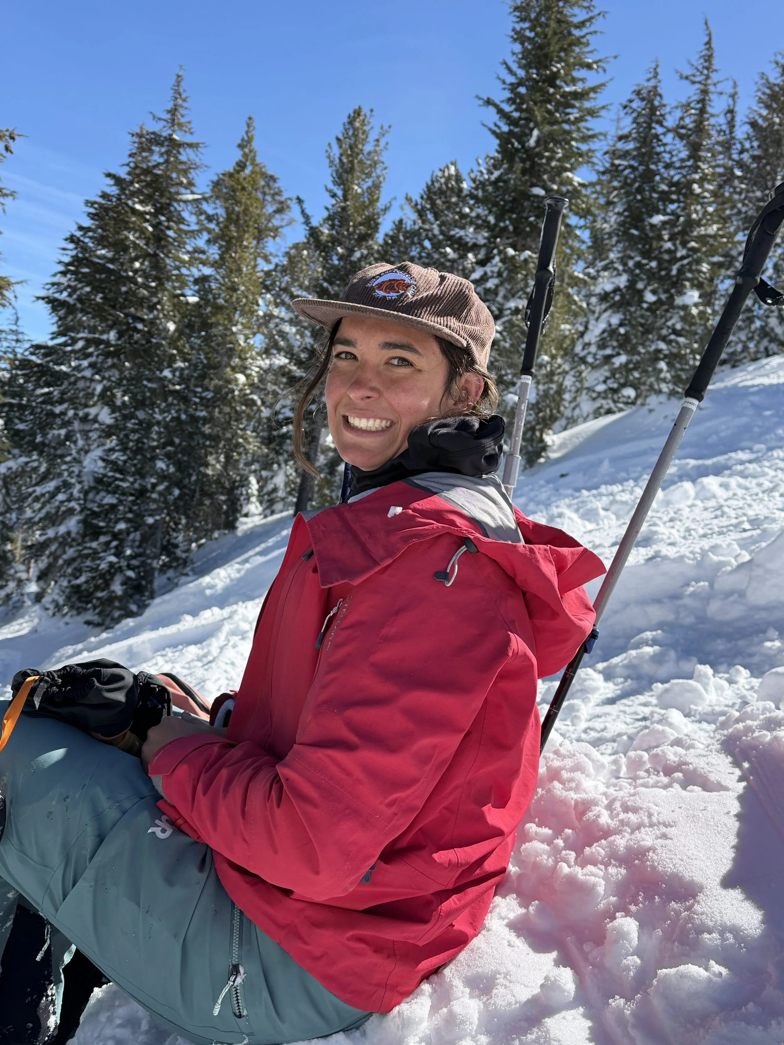 A smiling woman in a red jacket and gray pants sitting in the snow with snow-covered trees and a clear blue sky in the background during a sunny day.