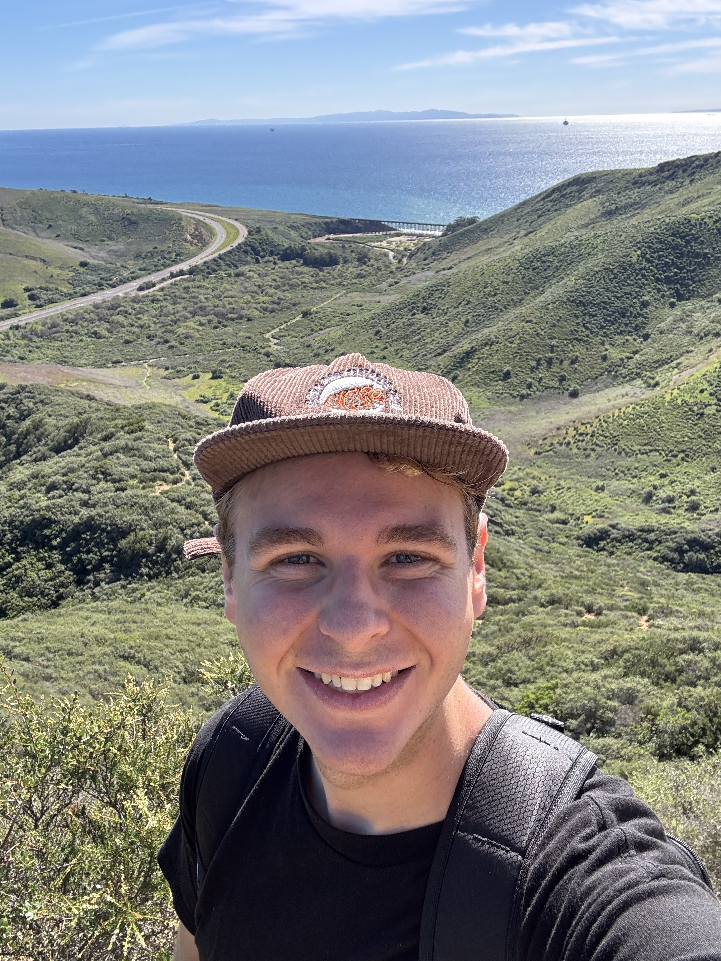 A young man smiling in front of a scenic coastal landscape with rolling green hills, a winding road, and the ocean in the background on a sunny day.