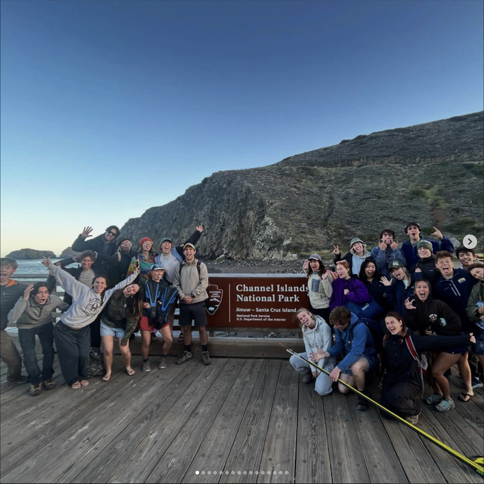 Group of people smiling and making gestures in front of a sign that says 'Channel Islands National Park, Santa Cruz Island, California' with a view of a mountain behind them.