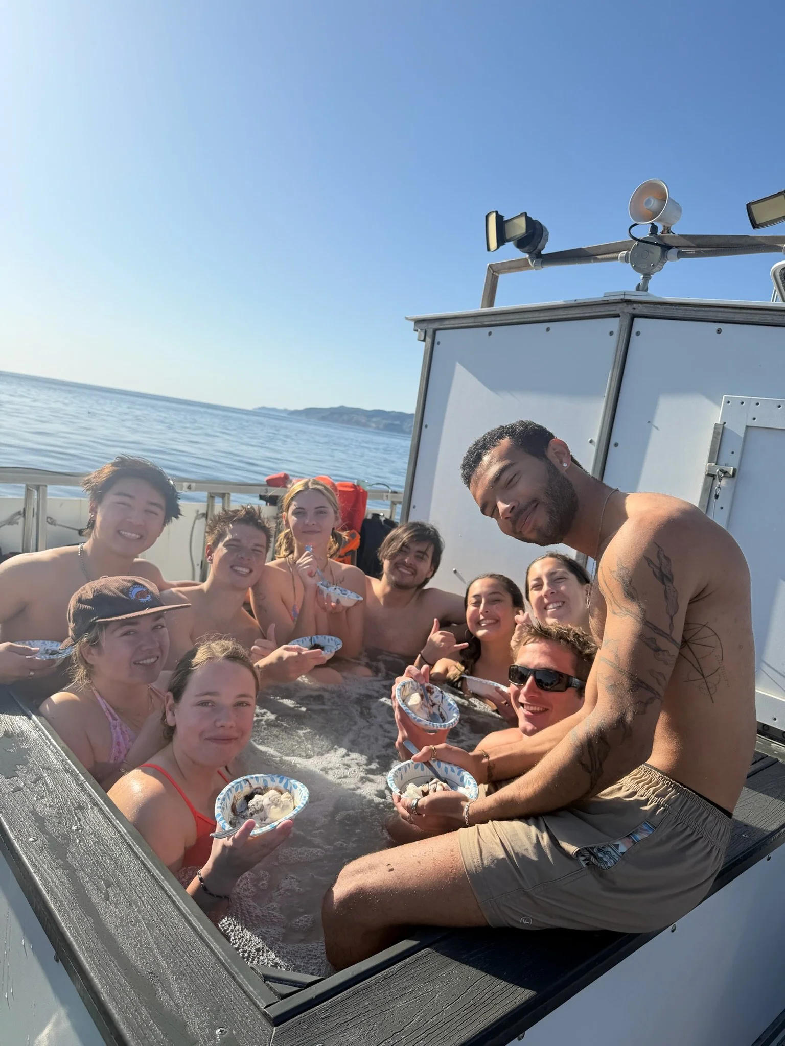 A group of people enjoying a hot tub on a boat with the ocean and distant shoreline in the background.