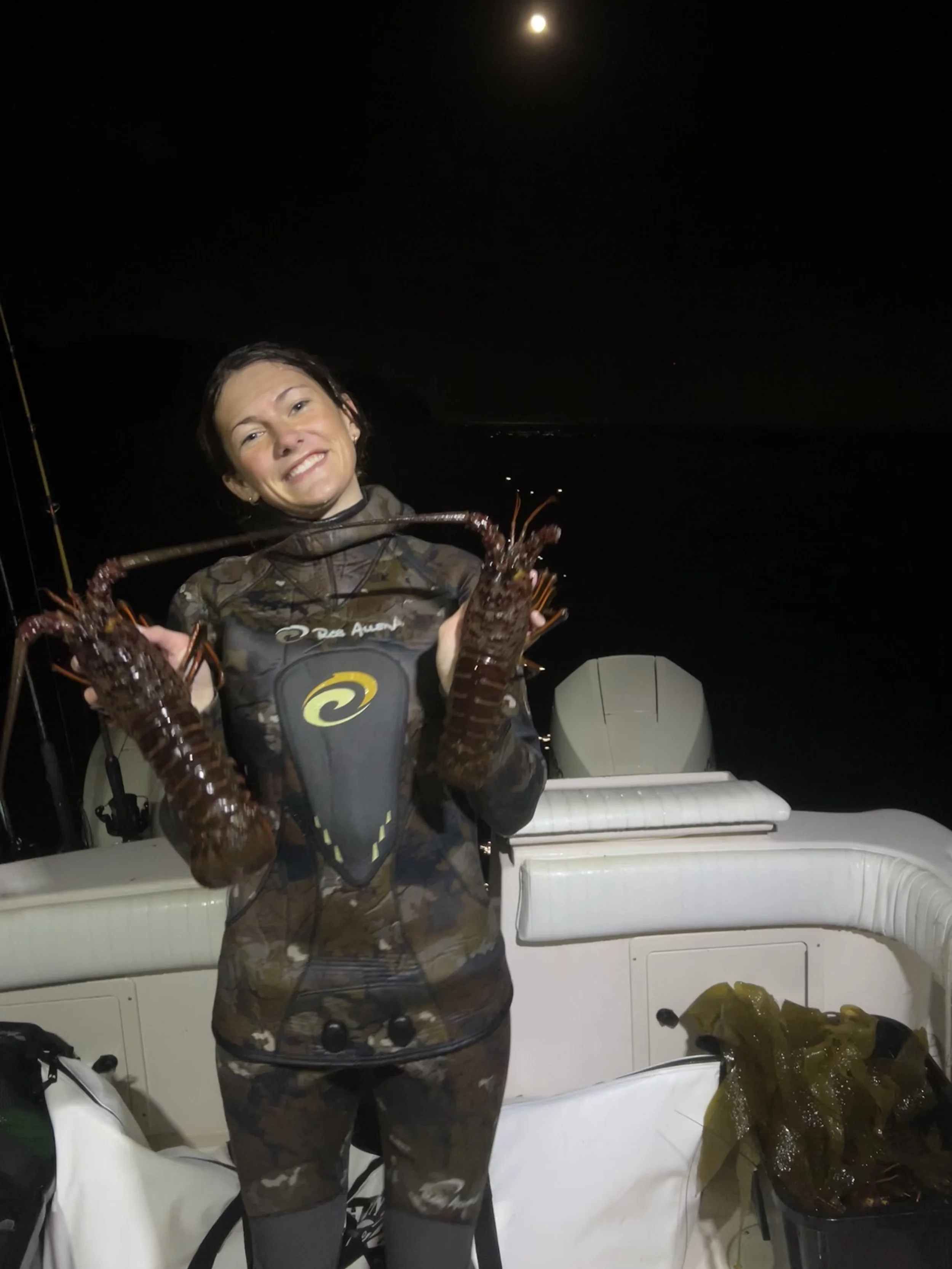 A woman in camouflage clothing smiling on a boat at night, holding two large lobsters.