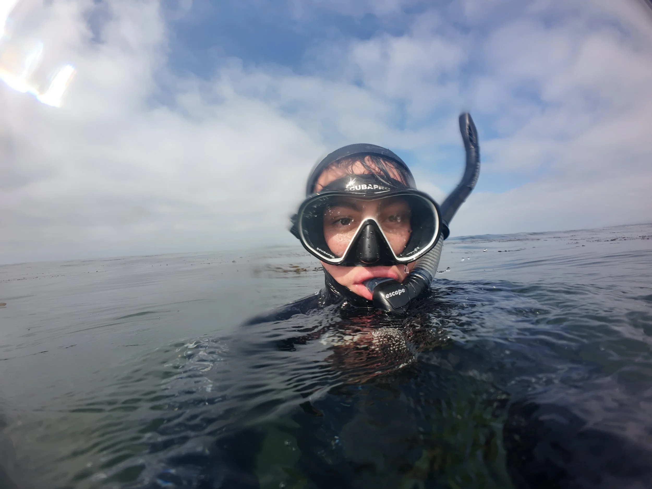 A person in a snorkel mask and wetsuit swimming in open water during daytime, with a cloudy sky in the background.