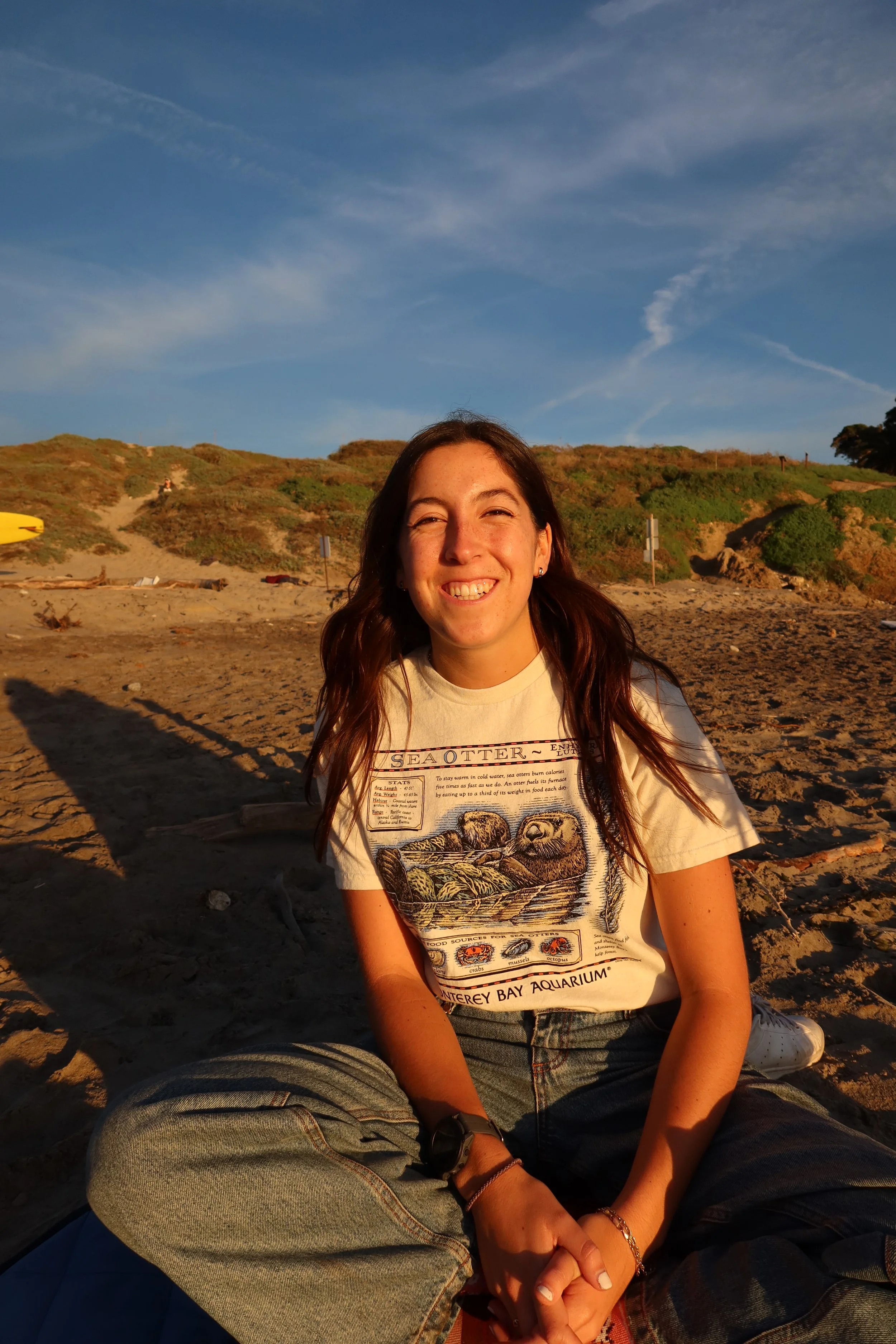 A woman sitting on a beach at sunset, smiling, with dunes and grassy hills in the background and a blue sky with wispy clouds.