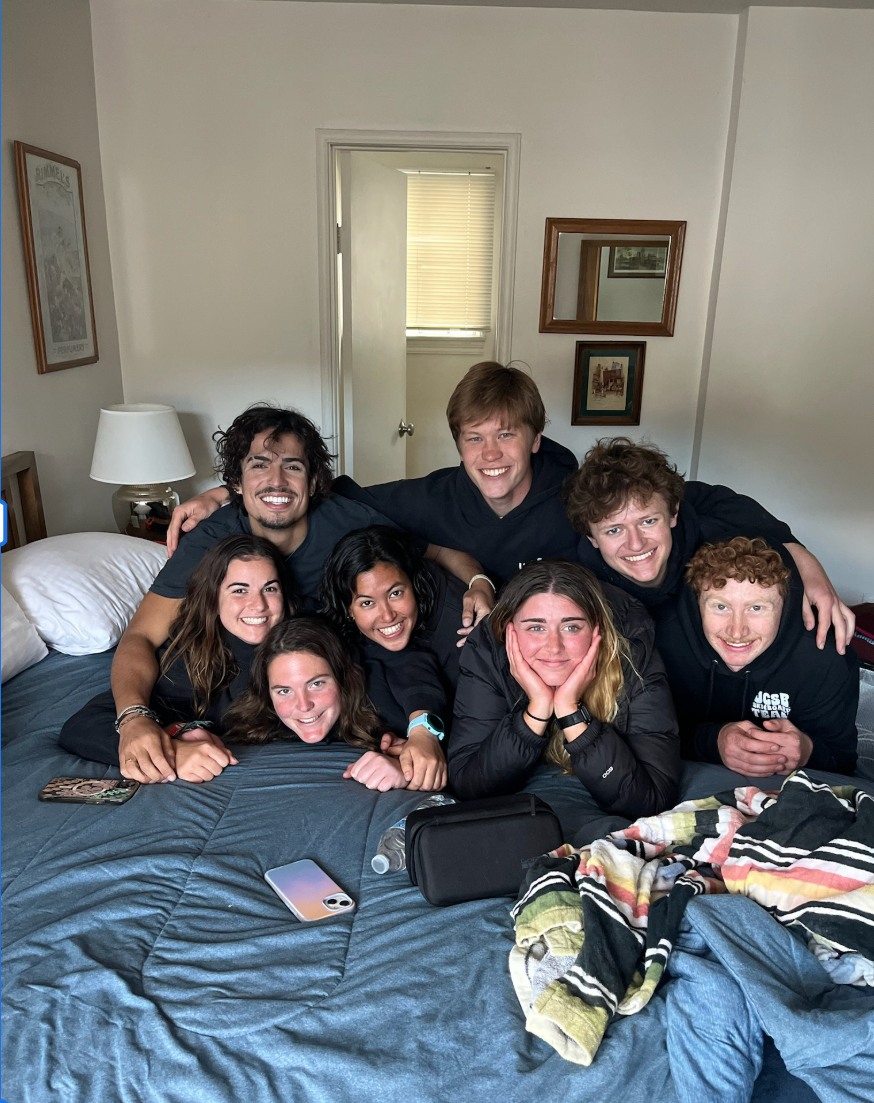 Eight young adults smiling and posing on a bed in a bedroom, with a lamp, framed pictures, and a closed door in the background.