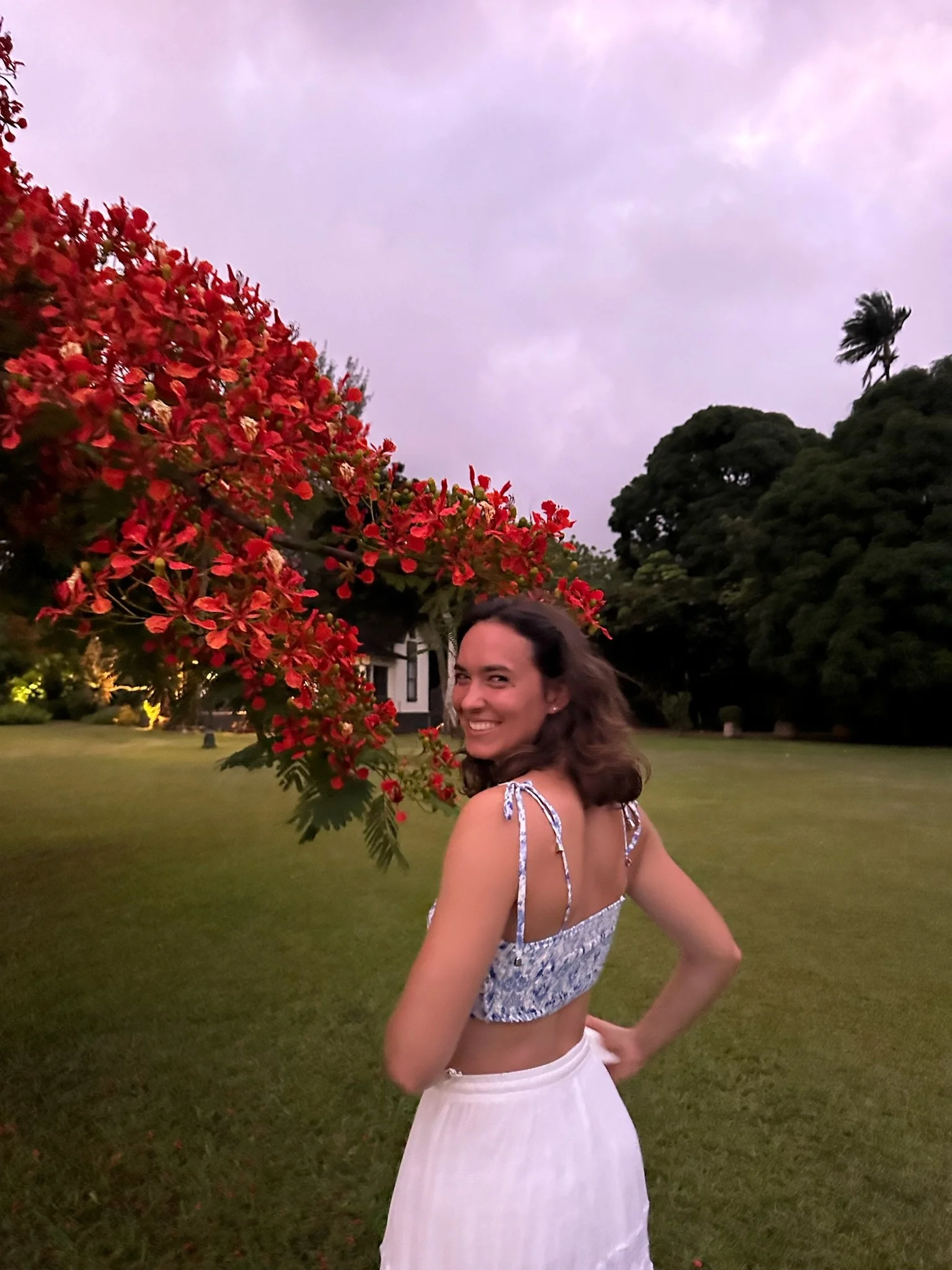A woman smiling and posing in a garden with red flowering tree, green grass, and large trees in the background during dusk or early evening.