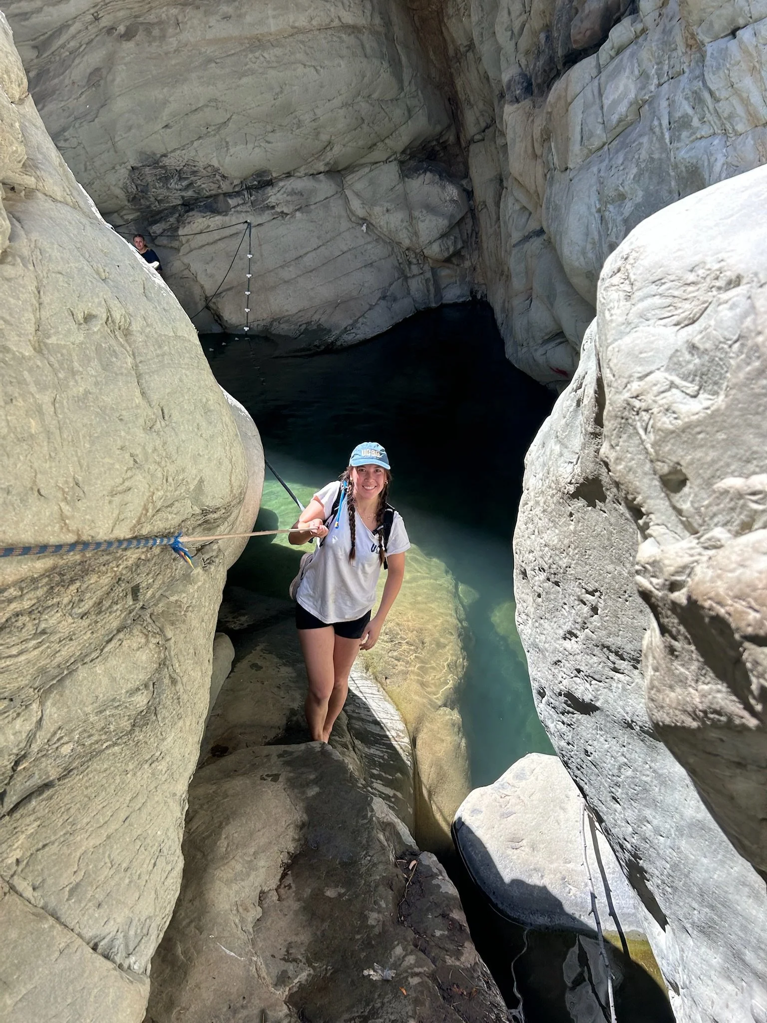 A woman is standing on rocks between canyon walls, holding a rope, with a girl seen in the background. They are near a body of water inside a narrow gorge.