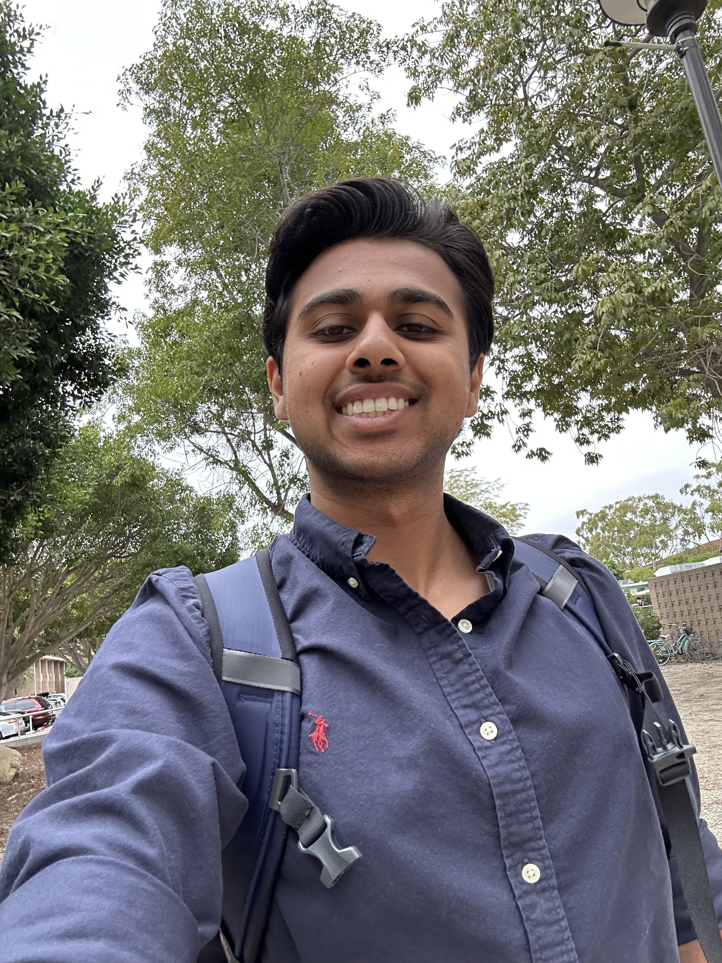 A young man taking a selfie outdoors, smiling, wearing a navy blue shirt with a backpack, with trees and parked cars in the background.